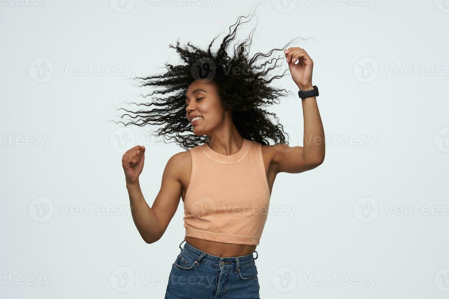 Cheerful joyful african american young woman with eyes closed and flying curly hair dancing and having party isolated over white background photo