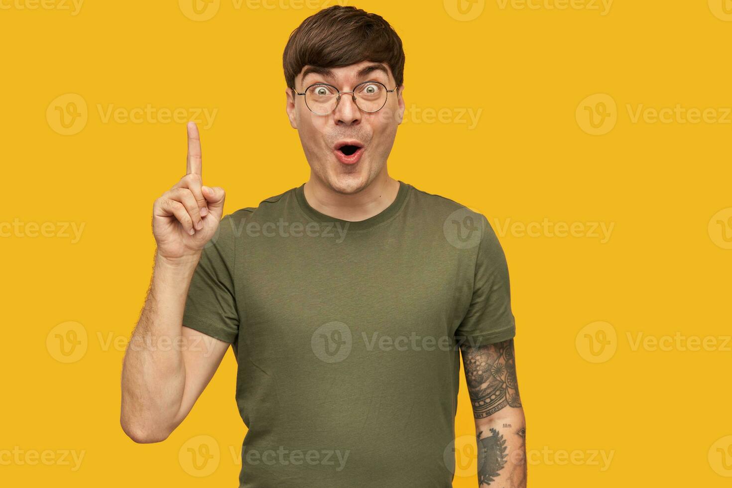 I have an idea Young attractive handsome male, wears green t-shirt, round glasses, point with a finger upwards, opened eyes widely, with amazed facial expression. Isolated over yellow background. photo