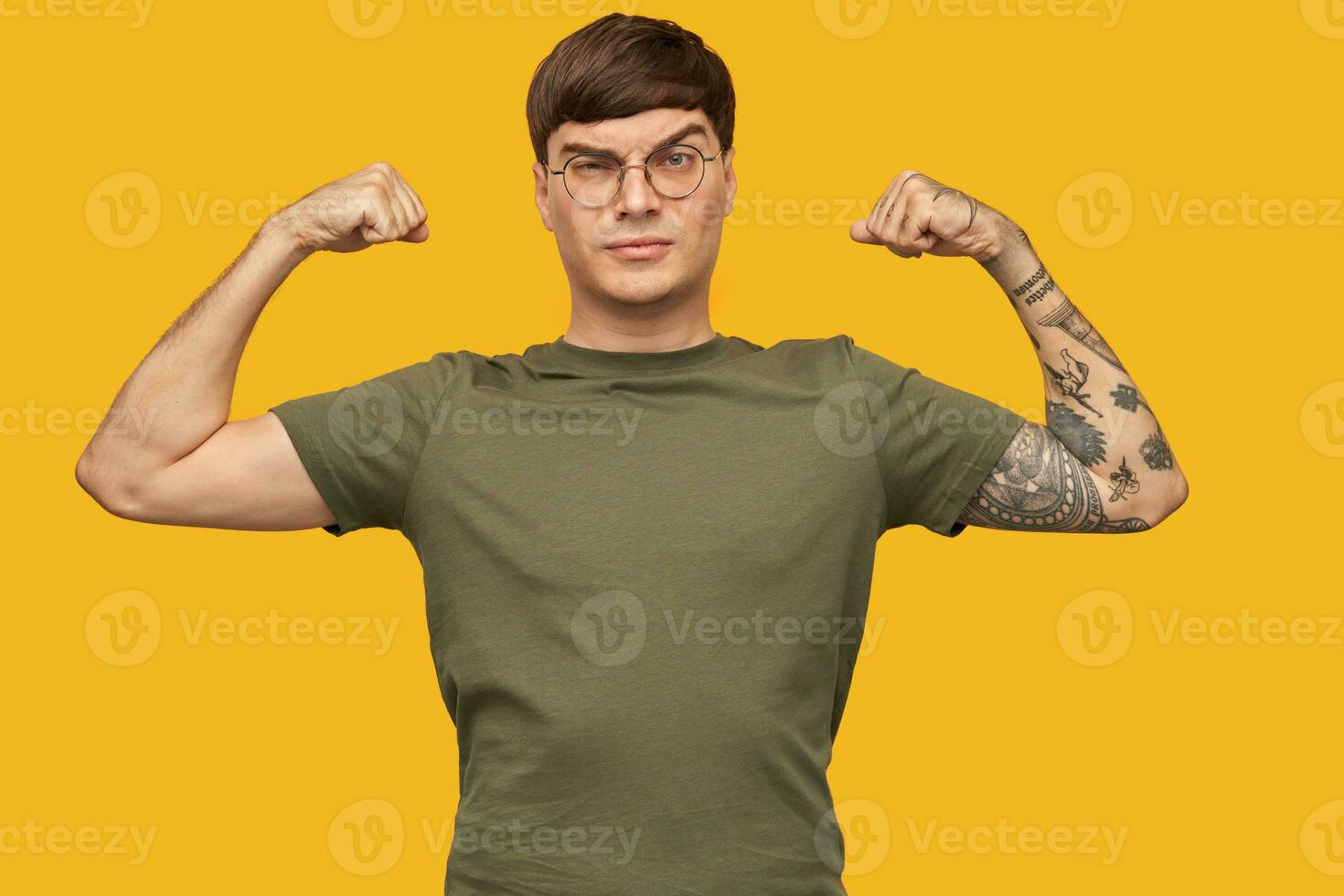 Indoor shot of serious young male, wears green t-shirt and round glasses, with tattooed hand, show his biceps and raised his eyebrow. Isolated over yellow background. photo