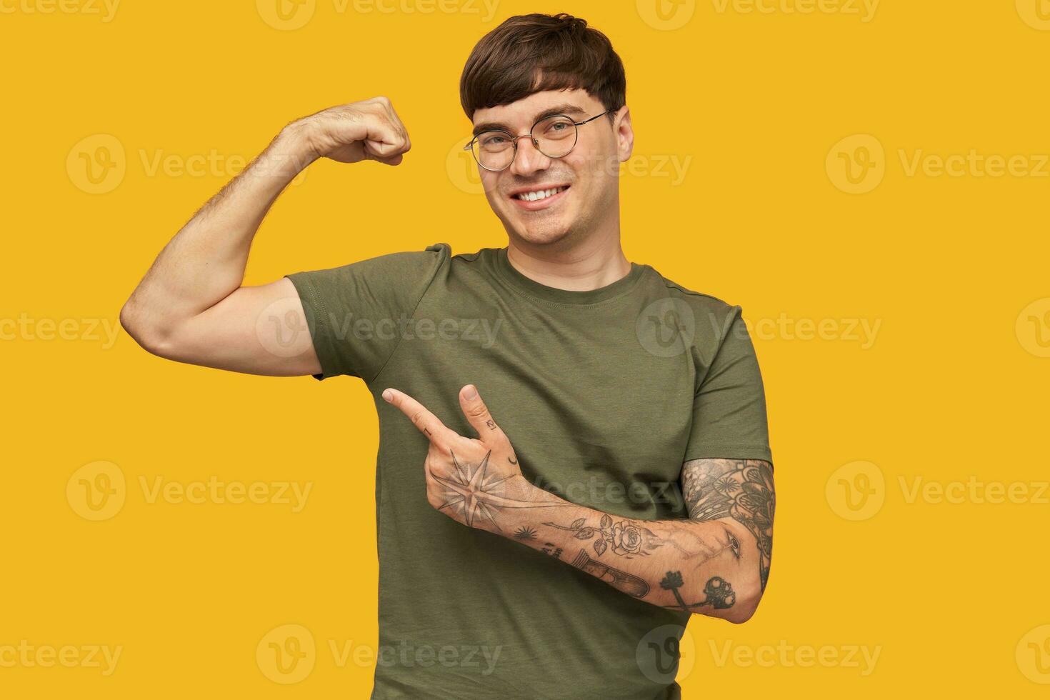 indoor shot of young attractive male, wears green t-shirt and round stylish glasses, indicates with a finger at his biceps and smiles broadly. Isolated over yellow background. photo