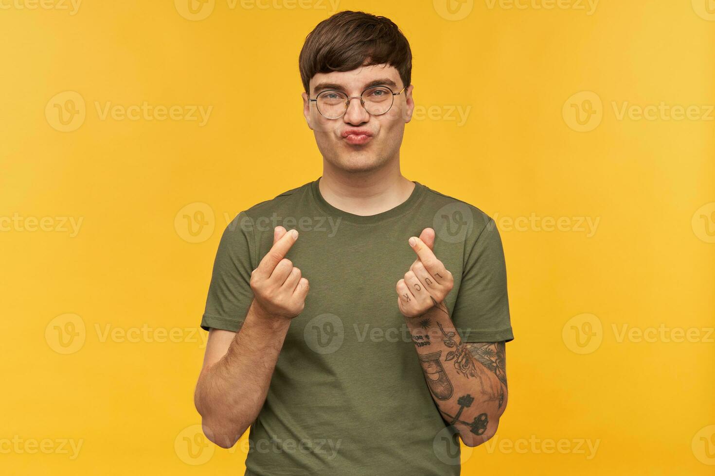 Indoor portrait of young funny male, shows money sign with both hands, curving his lips with happy facial expression. Isolated over yellow background. photo
