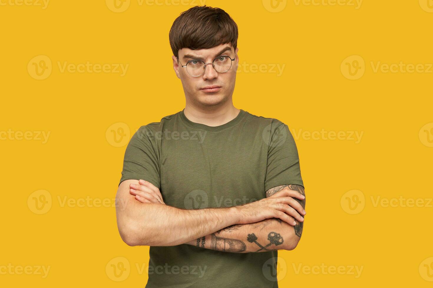 Indoor shot of young male looks seriously into camera with raised eyebrow, keeps his hand crossed on chest with confused, displeased facial expression. Isolated over yellow background. photo