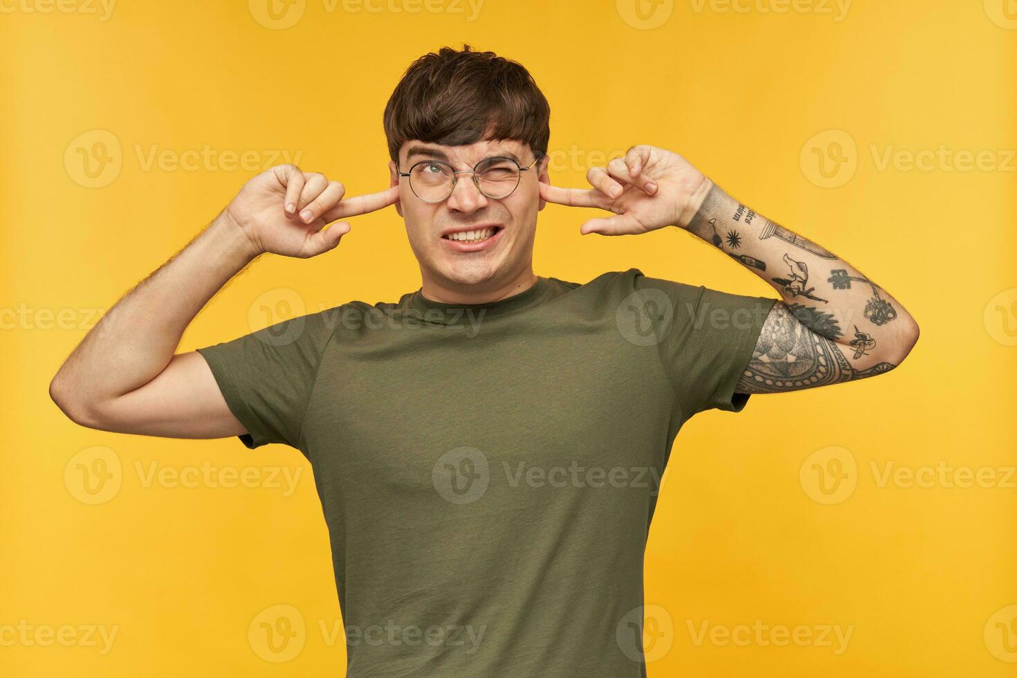 Indoor shot of young male student, looking upwards with negative, tired facial expression, plugs his ears with fingers because a lot of noise. Isolated over yellow background. photo