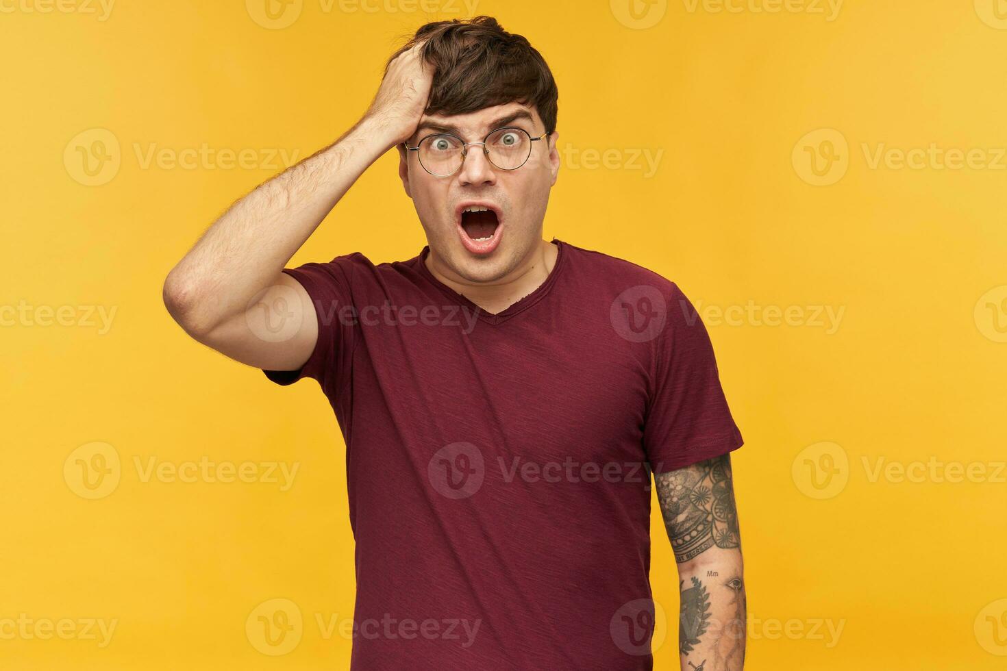 Indoor portrait of young male student wears dark red t-shirt, touching his head with hand, have a confused, negative facial expression. Isolated over yellow background. photo