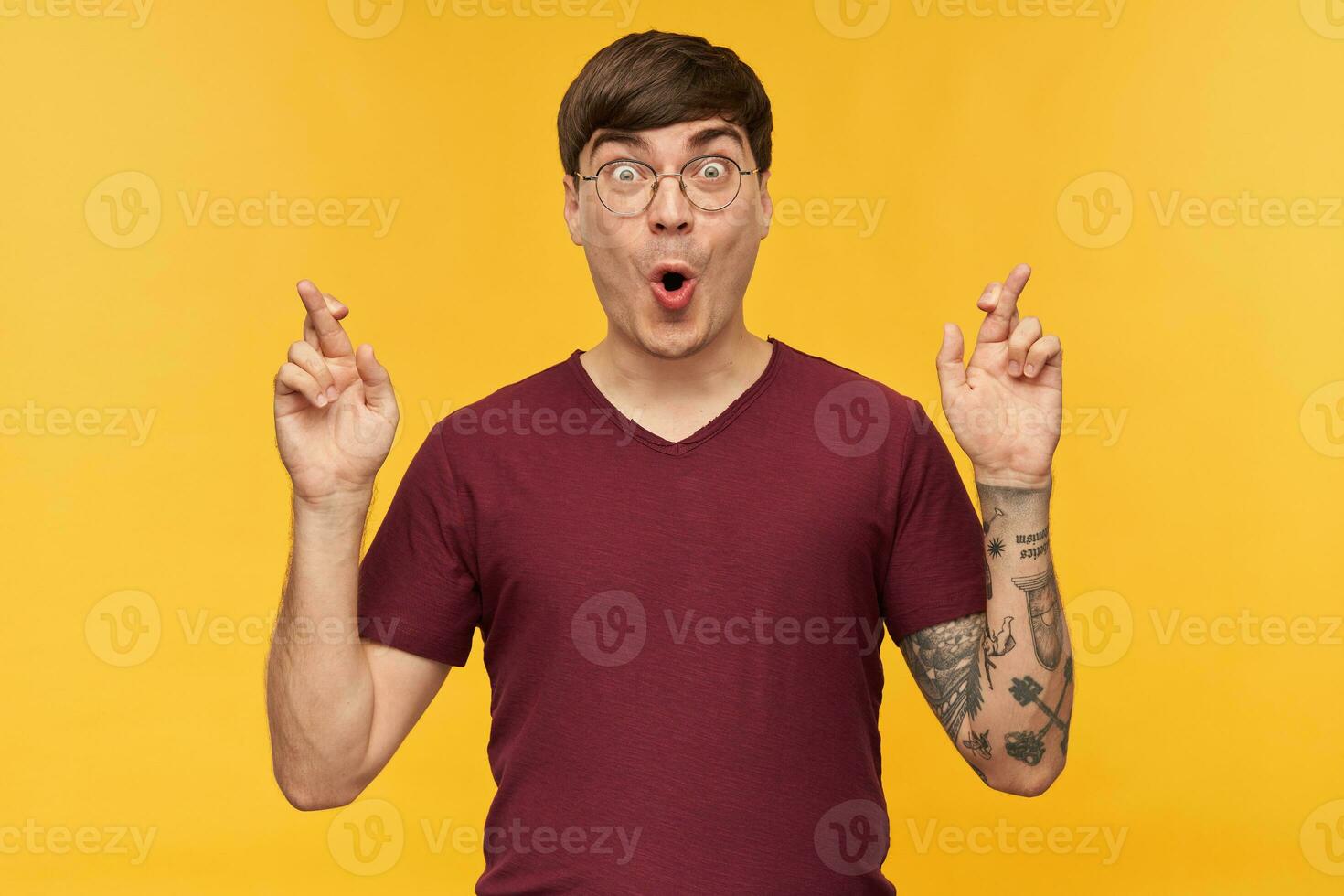 Indoor shot of young male student, keeps his finger crossed with amazed, shocked facial expression. Isolated over yellow studio background. photo