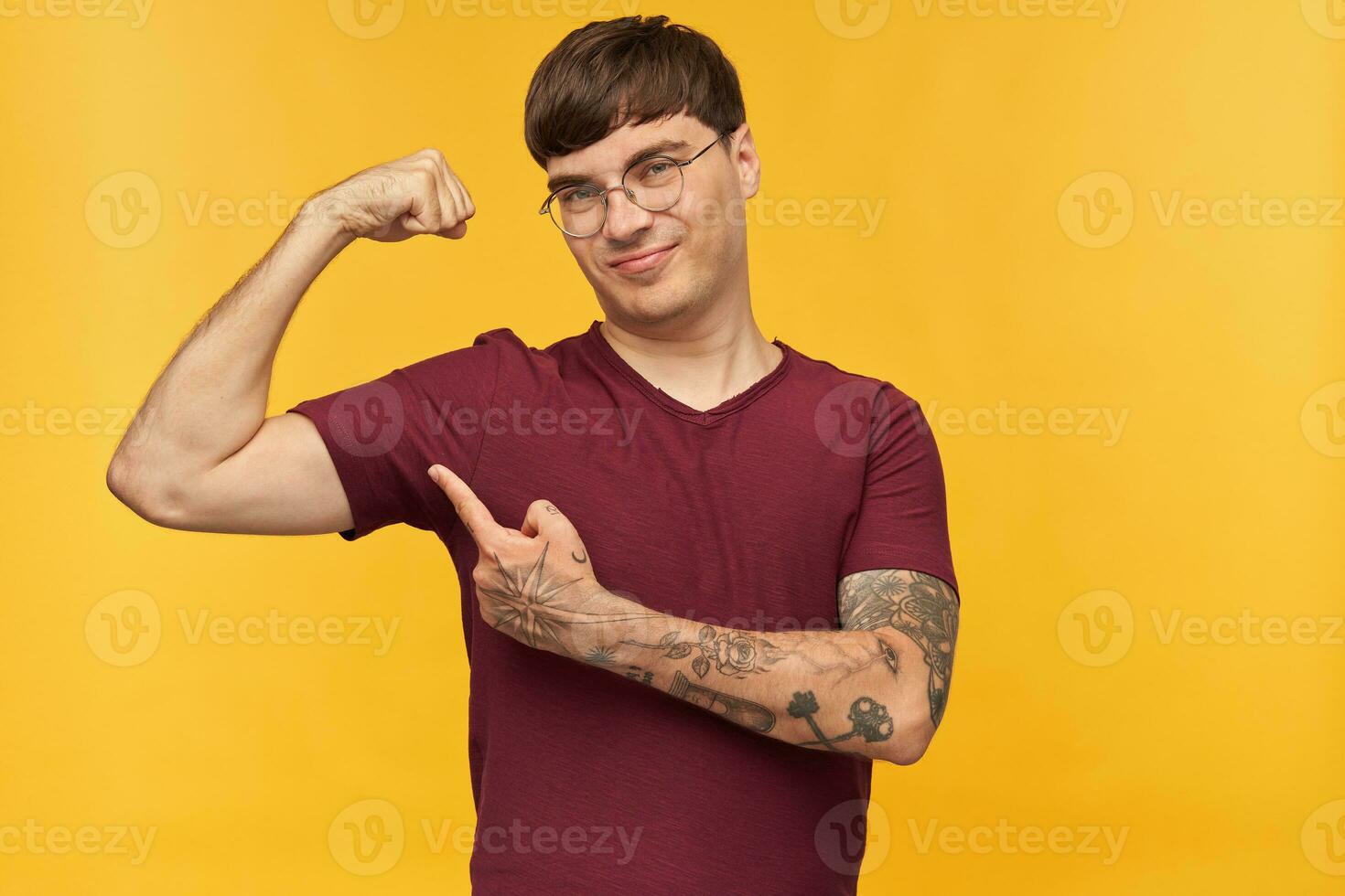 interior Disparo de joven masculino alumno, sonriente y señalando con un dedo a su bíceps, muestra cómo fuerte él es. aislado terminado amarillo antecedentes. foto