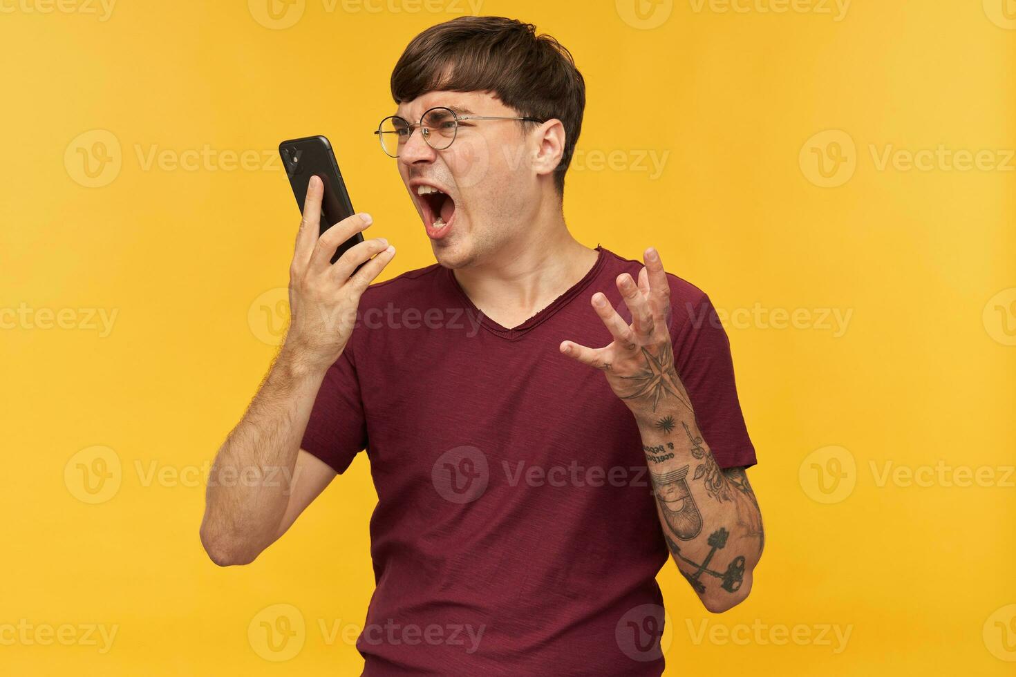 indoor shot of young negative, mad man, shouting into his phone while have a conversation, keeps his hand raised. Isolated over yellow background. photo