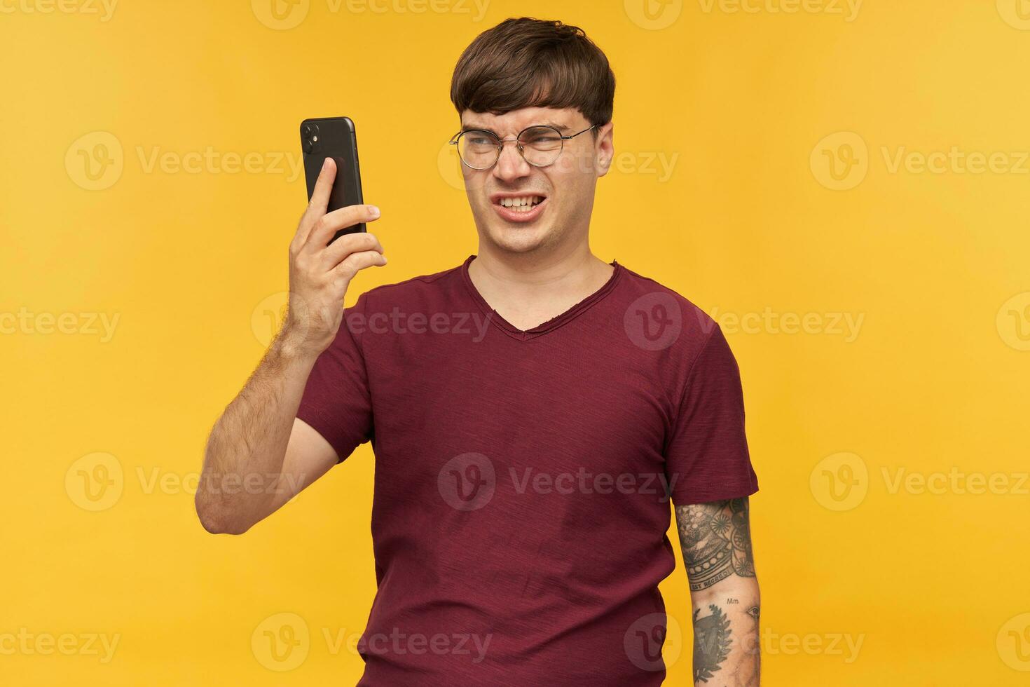 Irritated young male, wears red t-shirt and round glasses, looks into display of his phone with negative facial expression. Isolated over yellow background photo