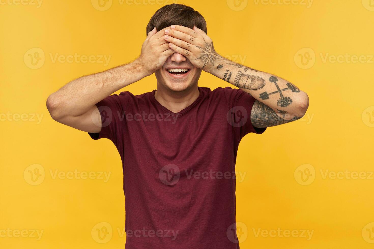 Indoor shot of young male wears red t-shirt, smiles broadly and closed his eyes with arms while waiting for gift. Isolated over yellow background. photo