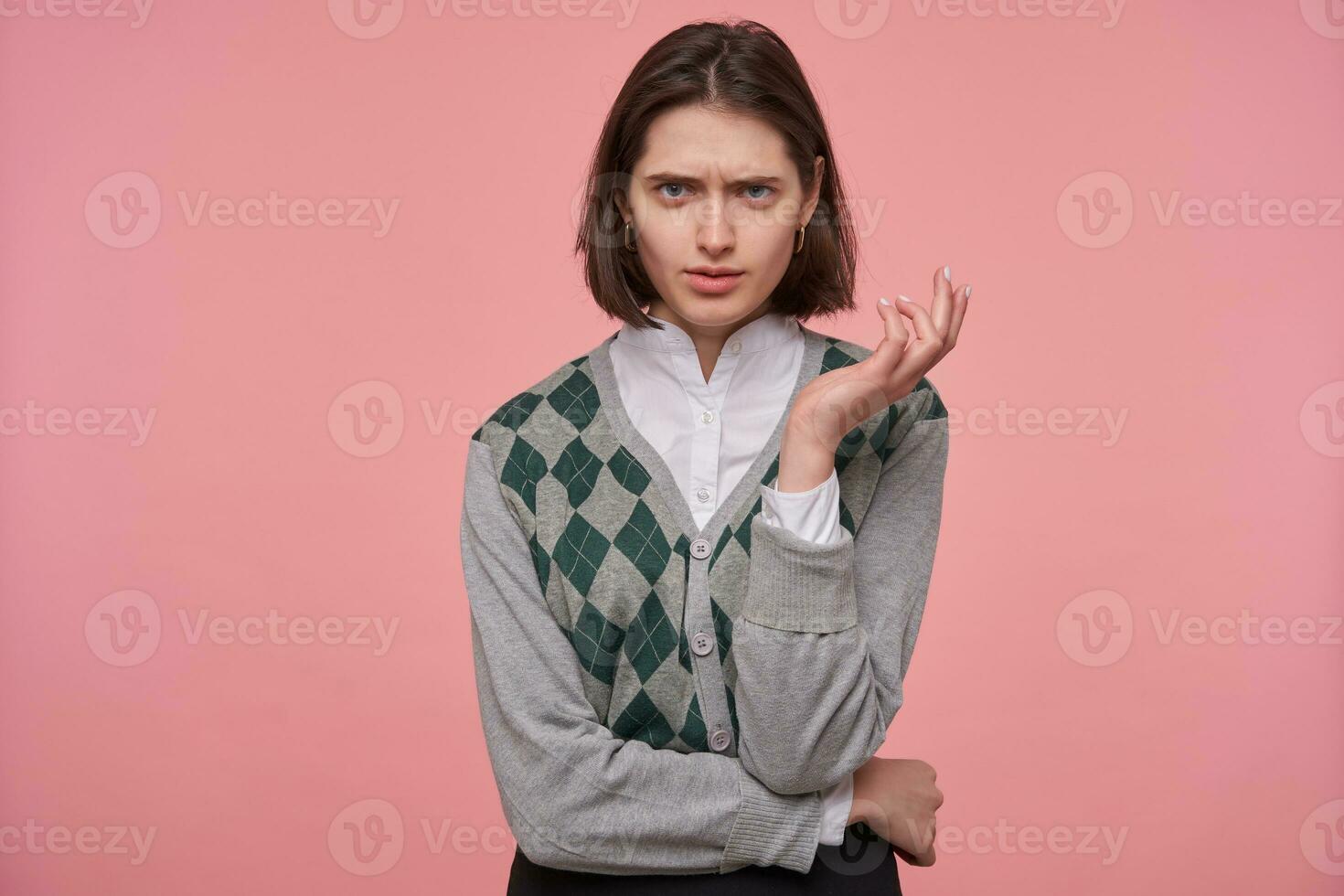 Indoor shot of young brunette student female, starring into camera with angry, mad facial expression, keeps her hand folded. Isolated over pink background. photo