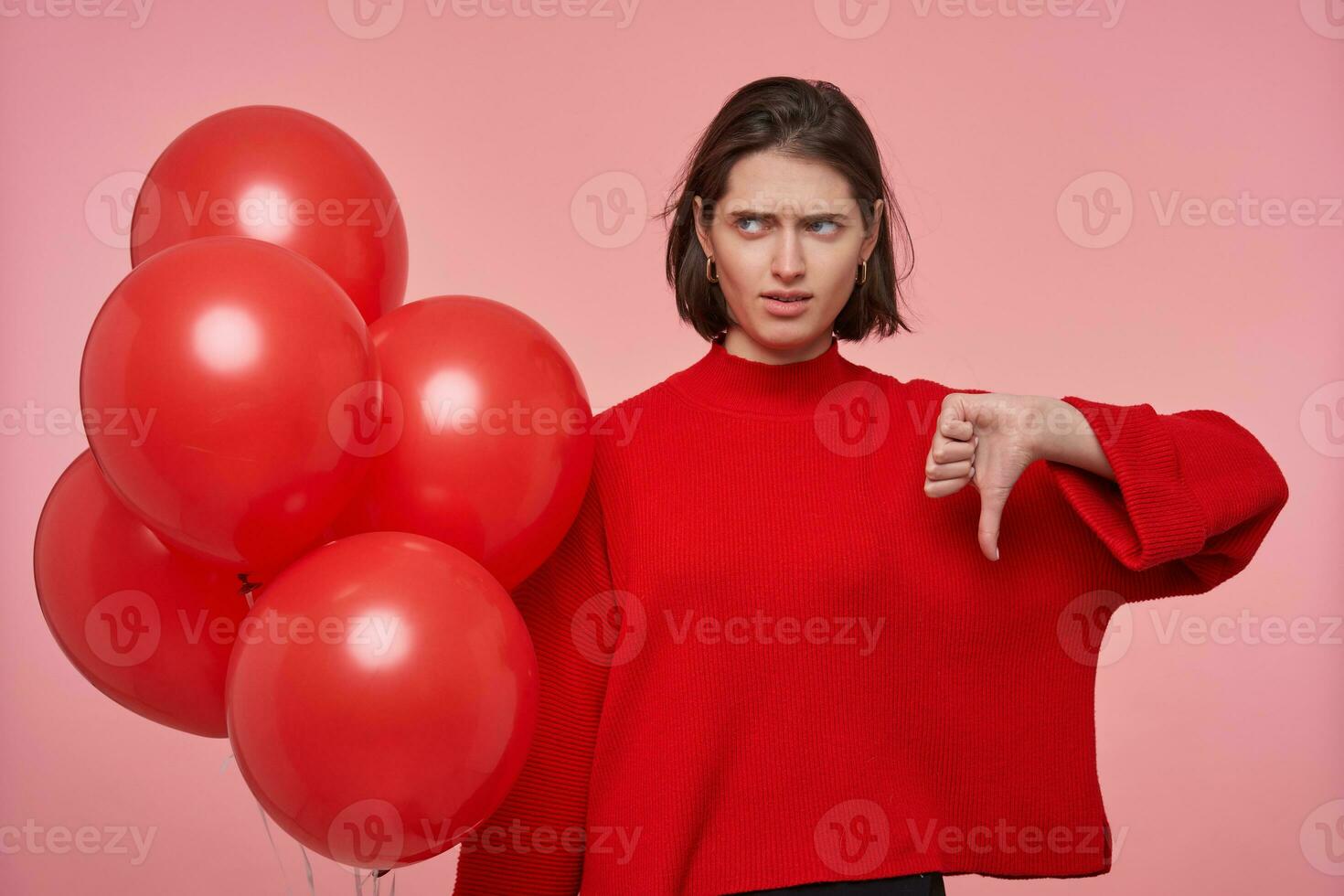Indoor portrait of young brunette female, wears bright red sweater golding balloons and shows thumbs down sign, fells unhappy, photo