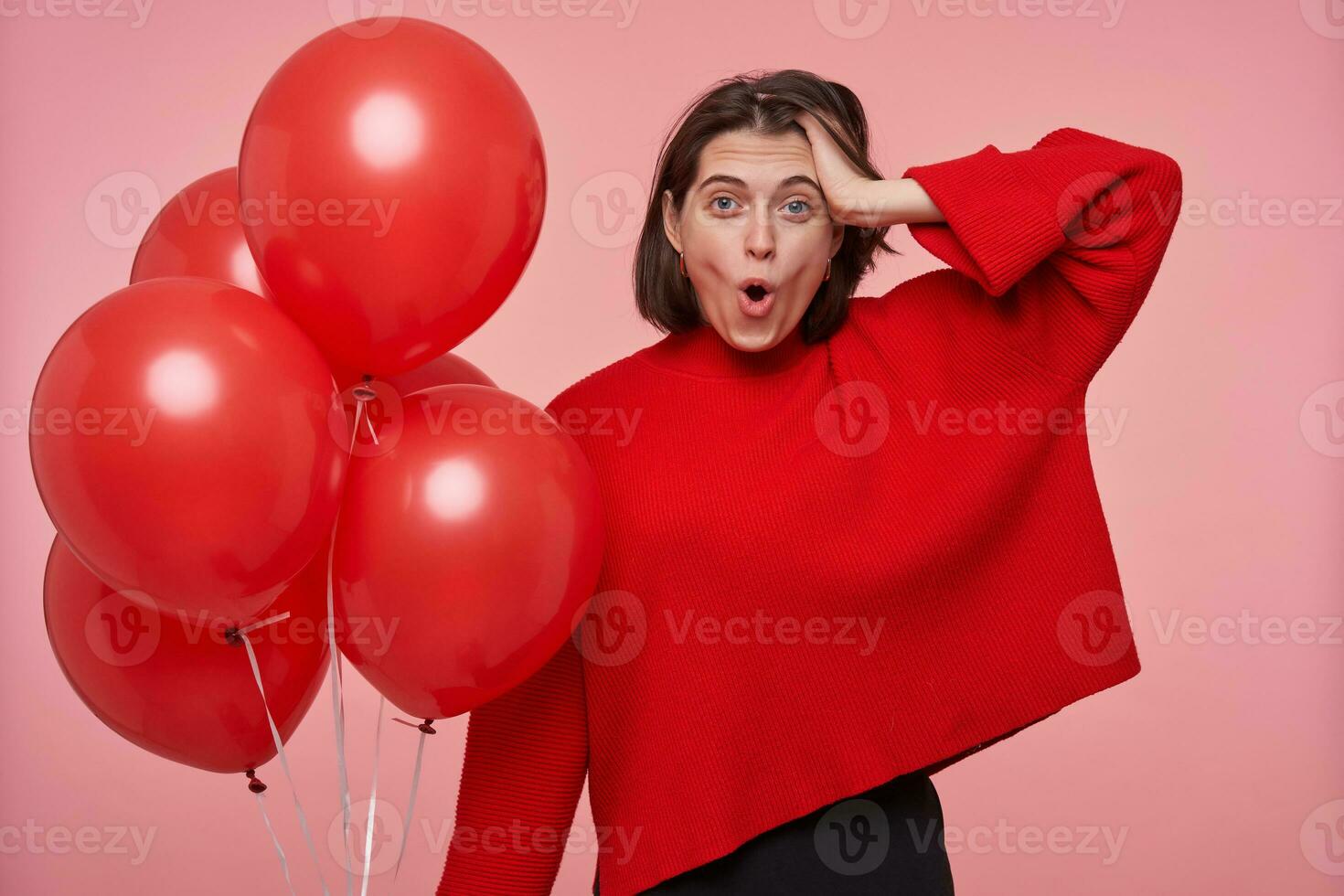 Young brunette female wears bright red sweater keeps mouth widely opened with shocked, surprised facial expression. Isolated over pink background photo