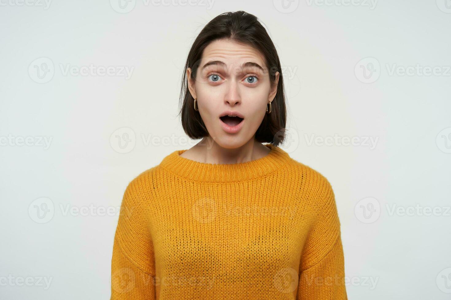 Indoor portrait of young brunette student female, feels confused and stressed, keeps her mouth widely opened with shocked facial expression, isolated over white background photo
