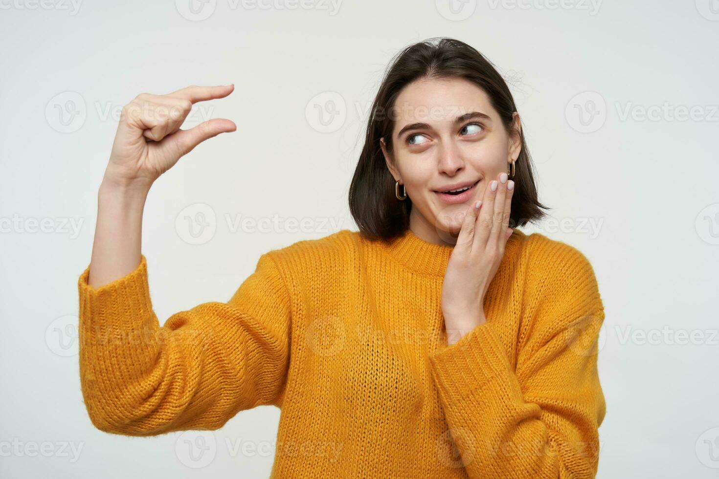 Indoor shot of young positive female wears yellow sweater, showing small gesture, smiles. Isolated over white background photo