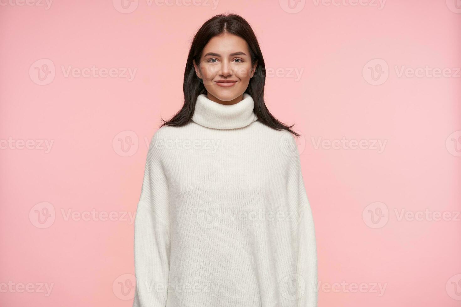 Indoor shot of young charming brown haired woman dressed in white wollen poloneck looking positively at camera with pleasant smile, standing over pink background photo