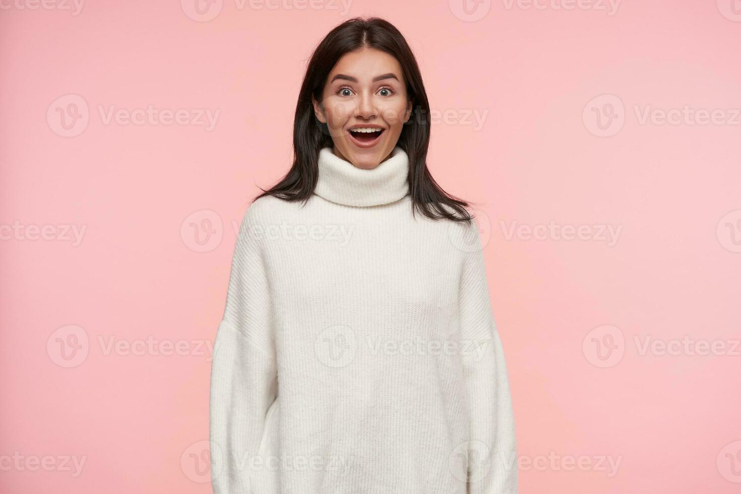 Surprised young lovely brunette female with loose hair looking excitedly at camera with opened mouth and keeping hands along body while posing over pink background photo