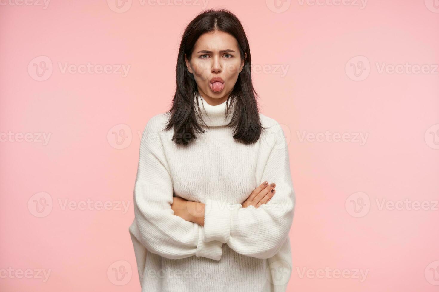 Offended young brown haired woman with casual hairstyle keeping her hands crossed and showing tongue to camera while standing against pink background photo