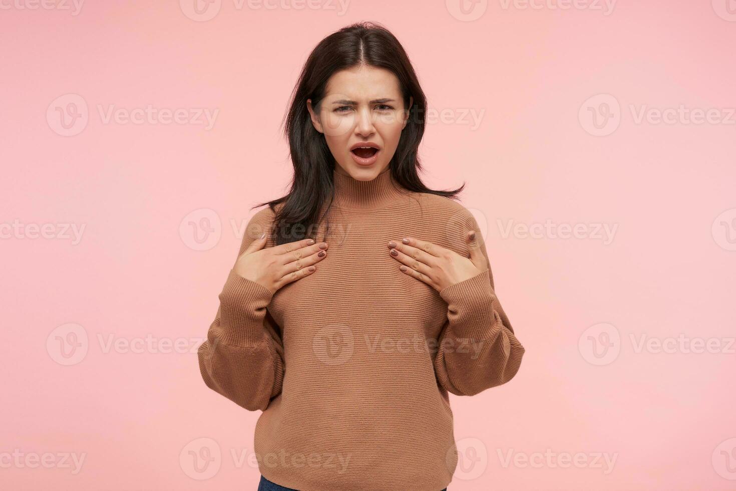 Puzzled young pretty brunette woman with casual hairstyle holding raised hands on her chest and squinting eyes while looking confusedly at camera, isolated over pink background photo