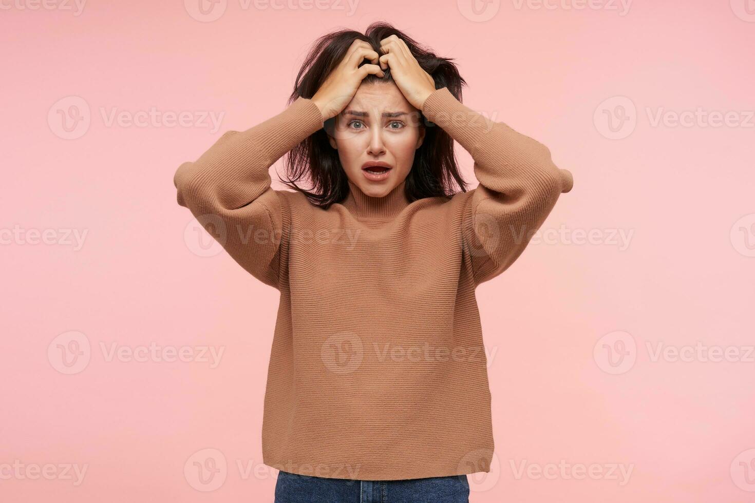 Bemused young bewildered brunette female with natural makeup holding her head with raised hands while looking confusedly at camera, isolated over pink background photo