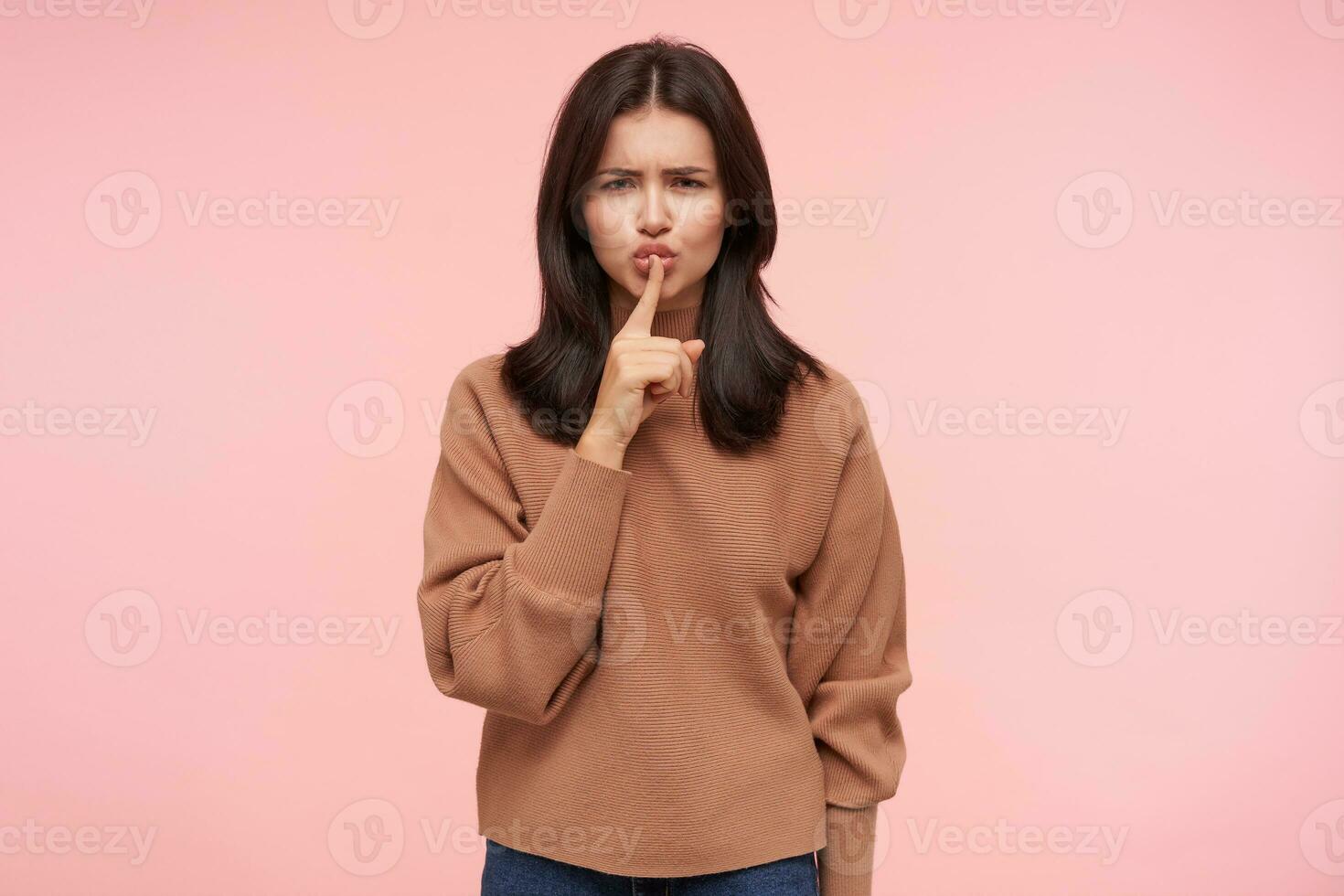 Severe young pretty brunette lady with casual hairstyle frowning her eyebrows while looking seriously at camera and holding index finger on her lips, isolated over pink background photo