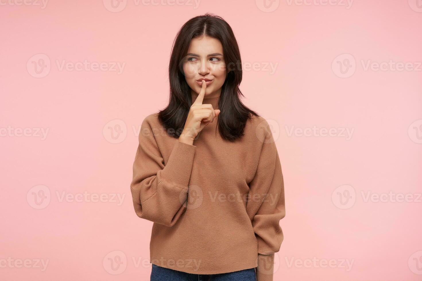 Glad young attractive brown haired woman with loose hair smiling cunningly while raising hand with hush gesture, standing over pink background in casual clothes photo