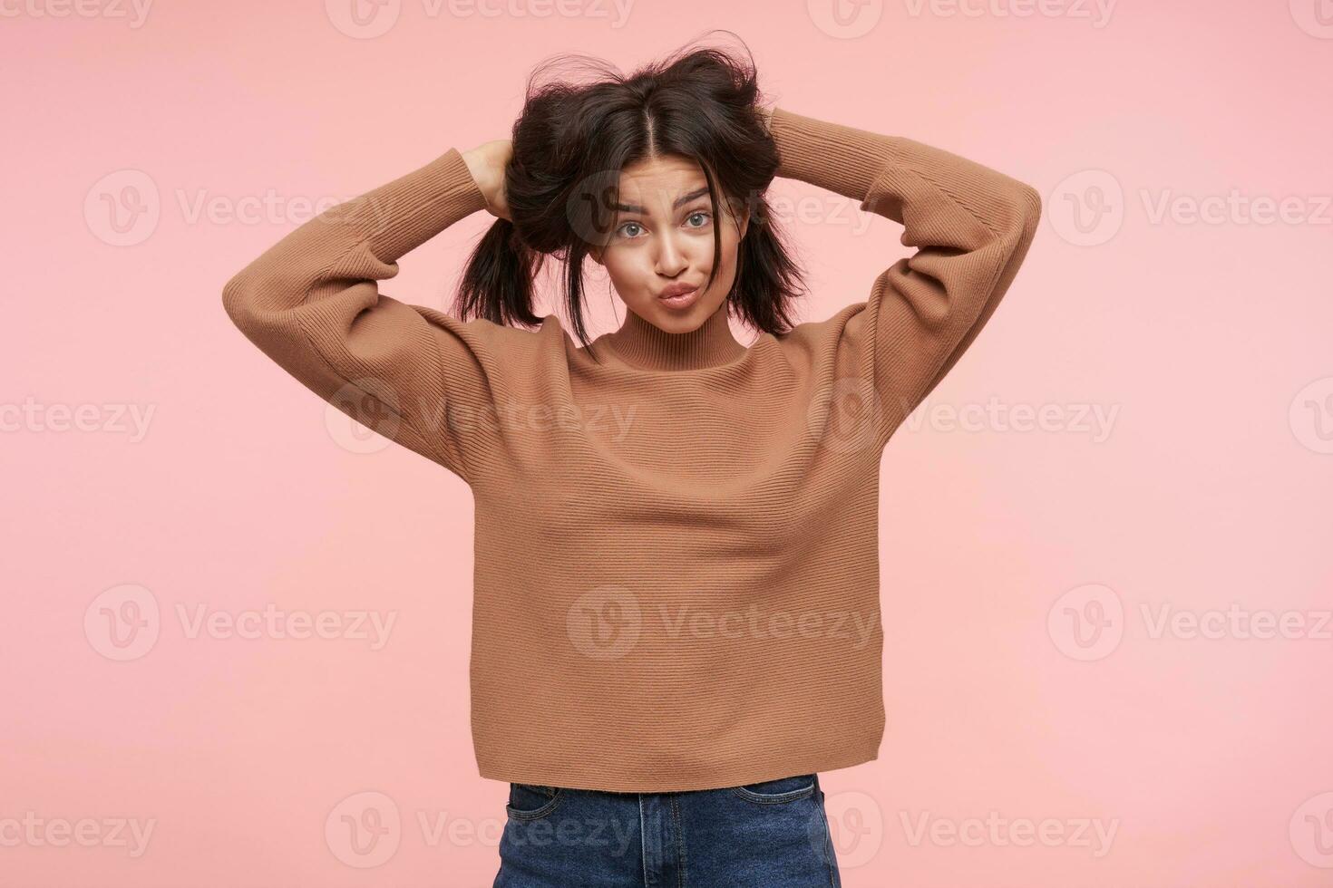 Studio photo of young funny brunette lady holding her hair with raised hands and pouting lips while looking at camera, standing over pink background