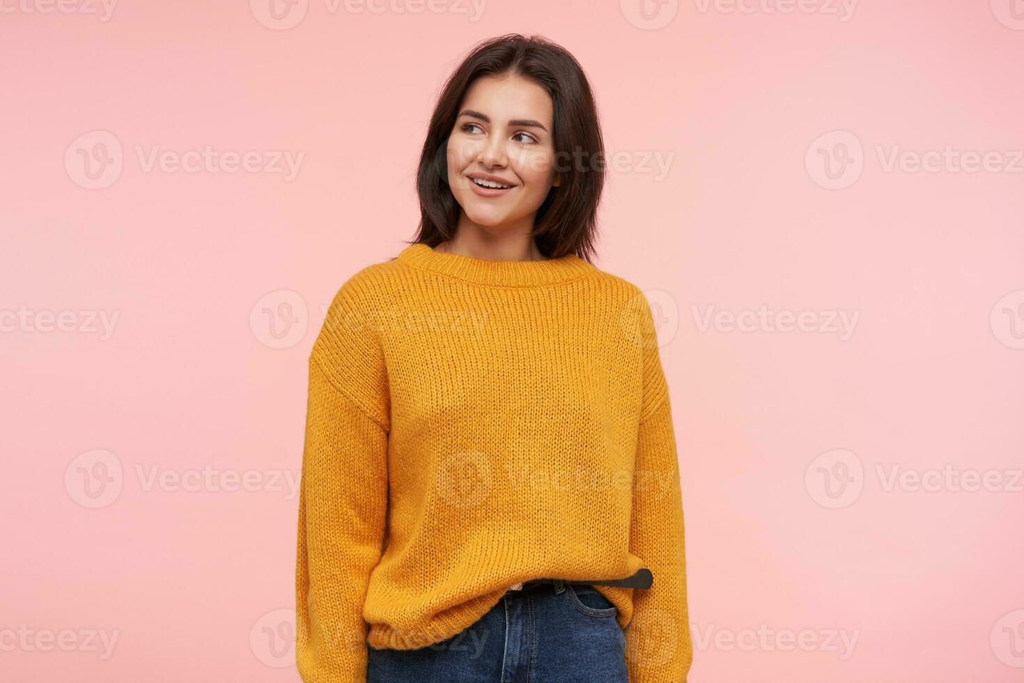 Cheerful young attractive brown haired woman with casual hairstyle smiling pleasantly and keeping hands along body while standing over pink background photo