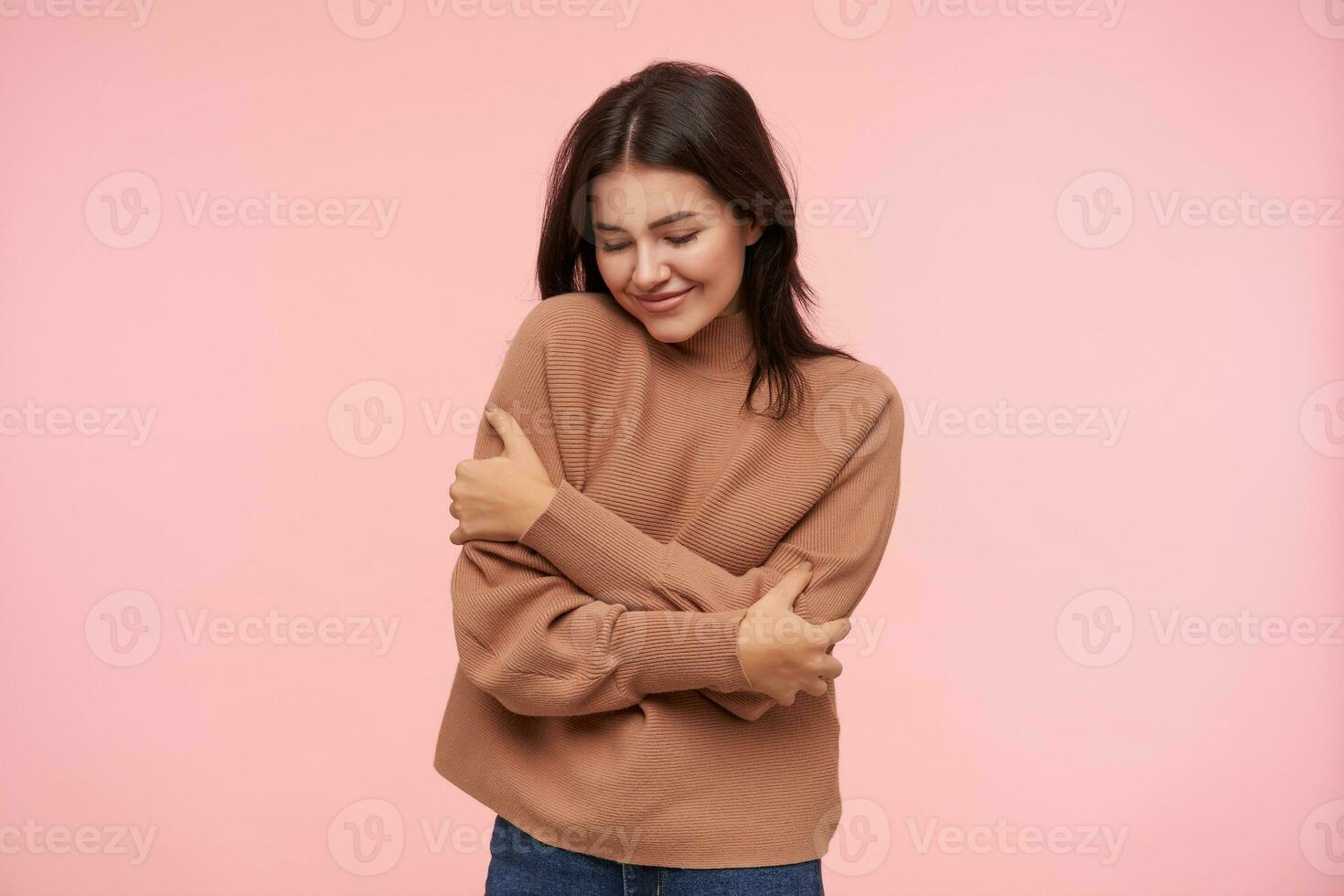 bueno mirando joven encantador marrón peludo mujer con casual peinado abrazando sí misma y sonriente bien mientras en pie terminado rosado antecedentes en marrón de punto suéter foto