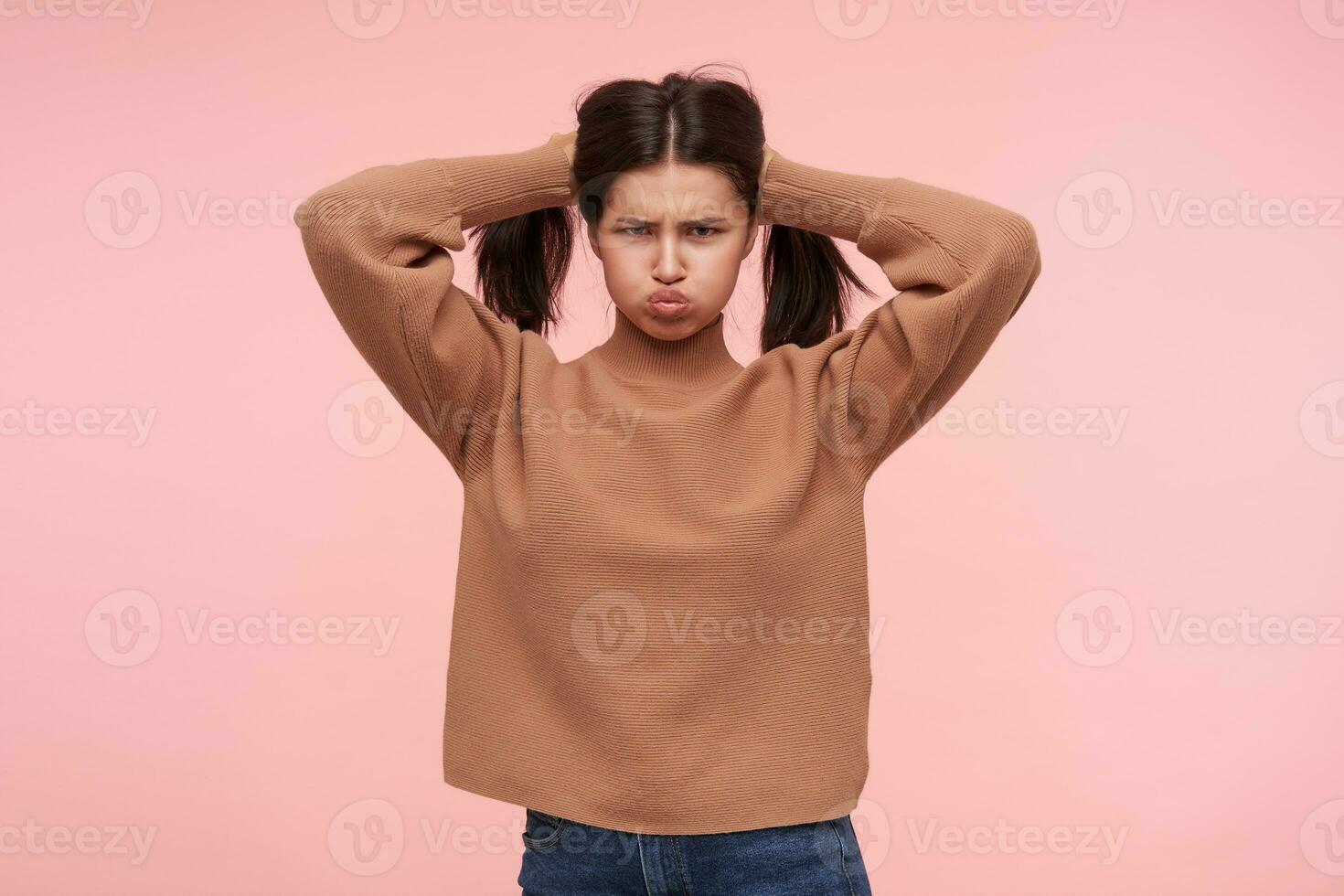 Gloomy young brown haired woman holding her hair with raised hands and puffing out her cheeks while looking severely at camera, standing over pink background photo