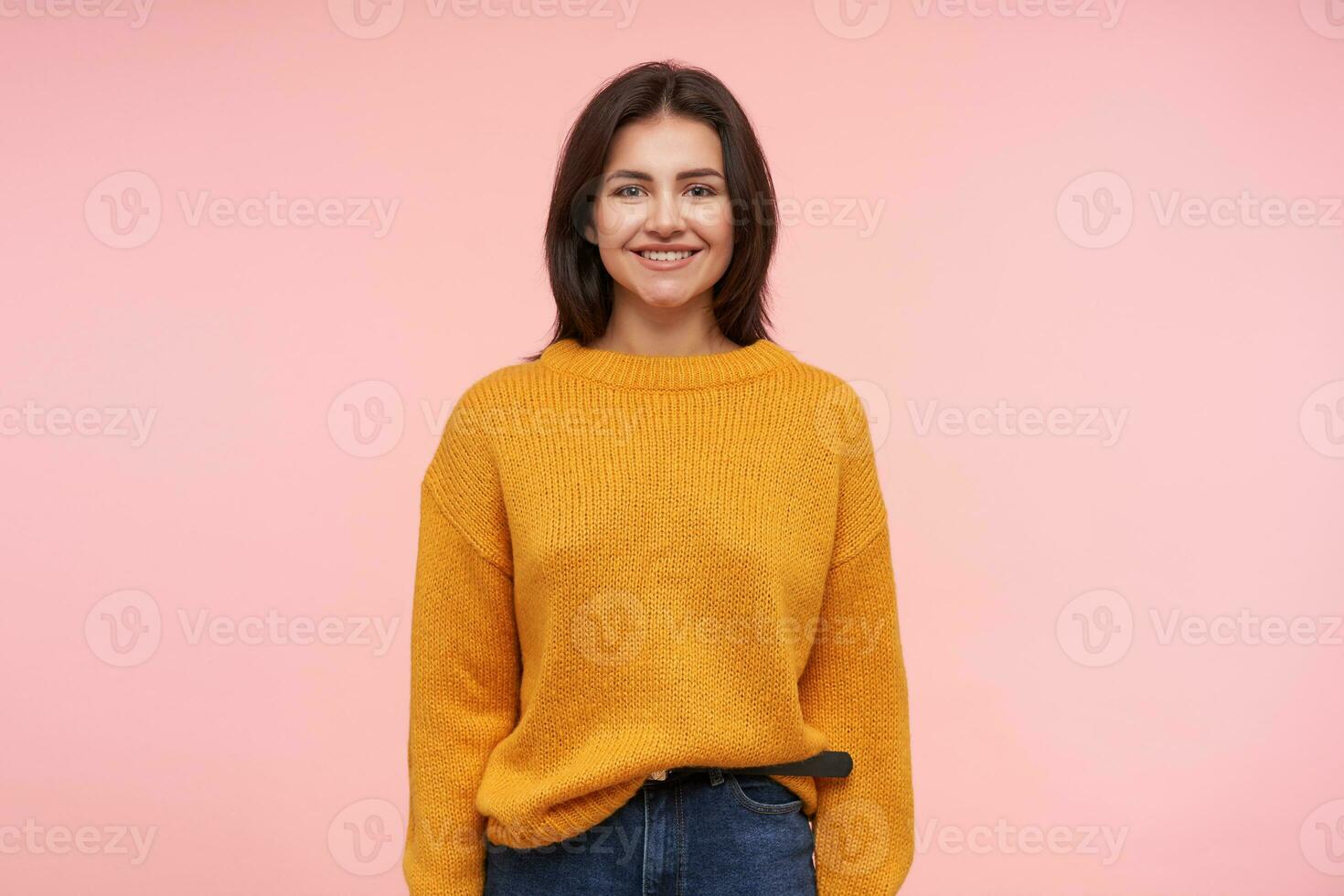 Pleased young attractive brunette female showing her white perfect teeth while smiling gladly to camera, standing over pink background with hands down photo