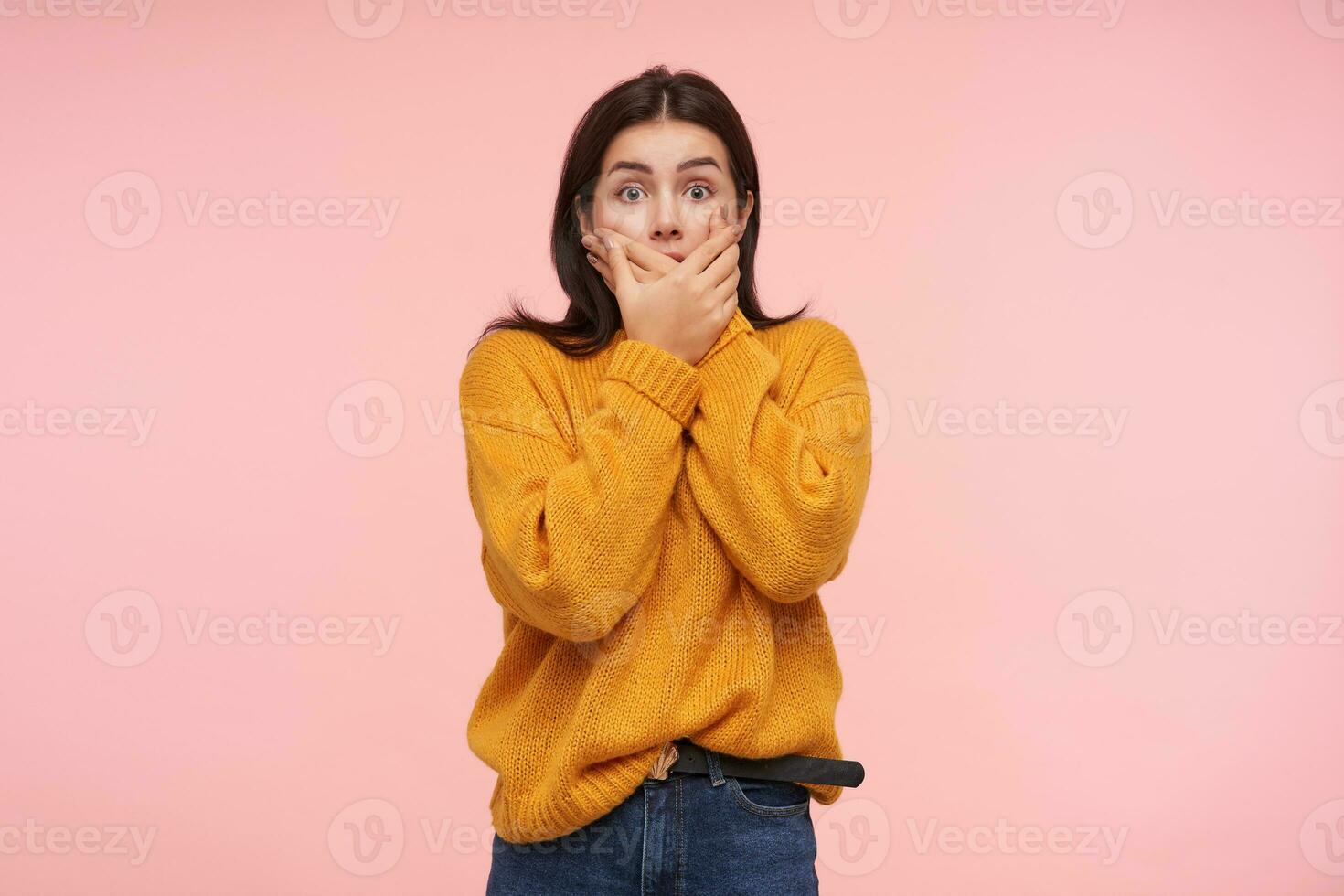 Studio photo of young shocked brunette lady with casual hairstyle covering her mouth with raised palms and looking amazedly at camera with wide eyes opened, posing over pink background