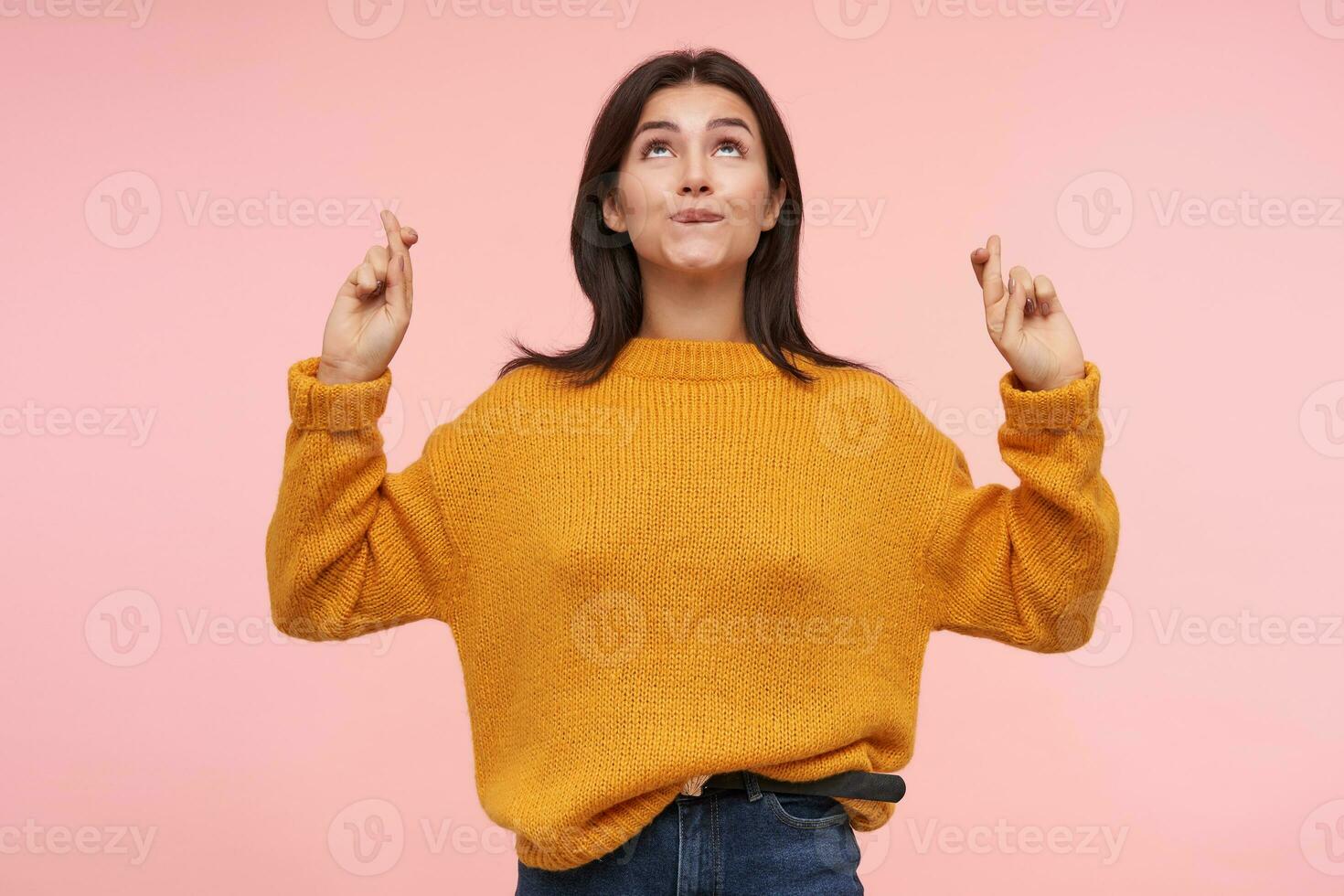 Positive young lovely brown haired female biting worringly underlip while looking upwards and raising hands with crossed fingers, standing over pink background photo