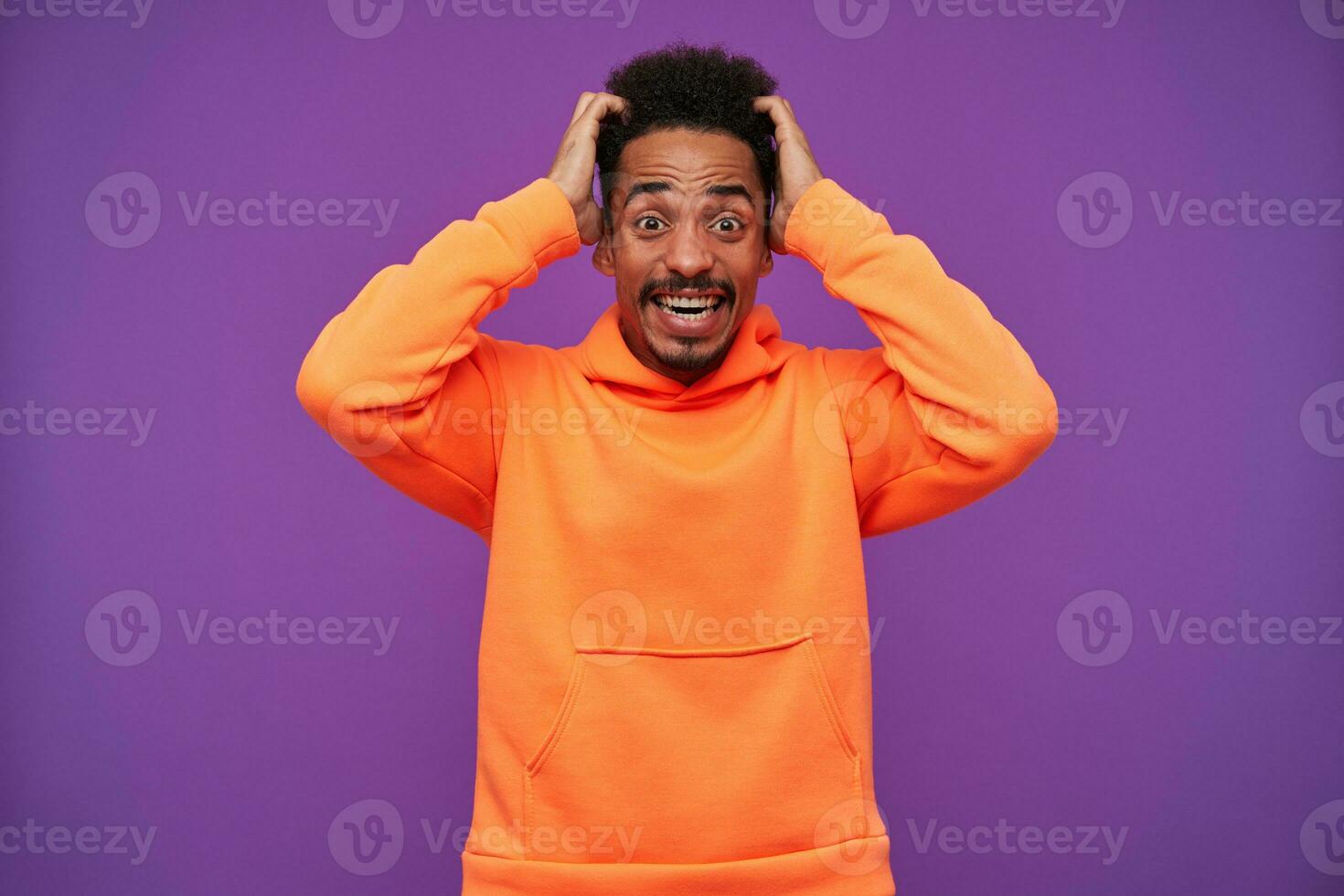 Overwhelmed young dark haired bearded dark skinned guy rounding his brown eyes while looking shocked at camera, holding his head with raised hands while posing over purple background photo