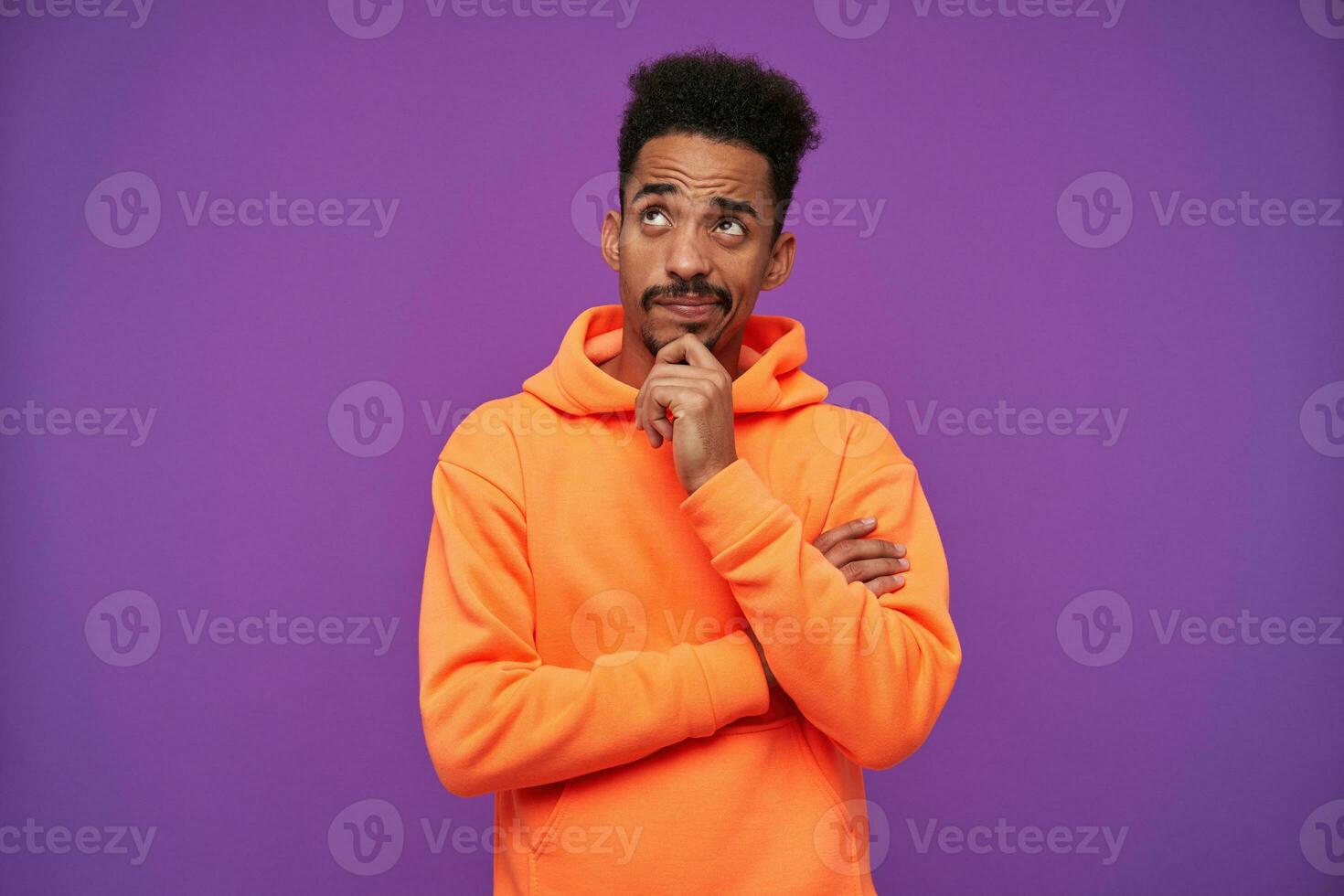 Pensive young attractive brown-eyed bearded brunette man with dark skin holding his chin with raised hand and looking thoughtfully upwards, wearing orange hoodie over purple background photo