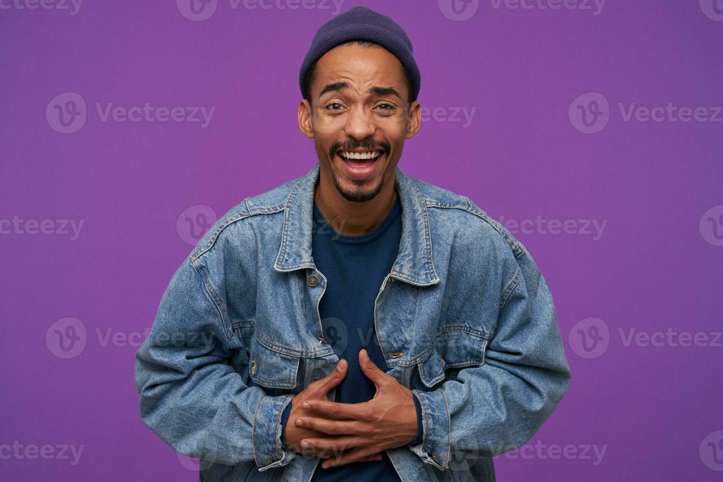 Indoor photo of young attractive dark skinned brunette male with beard cluthing at his belly with hands while laughing joyfully while looking happily at camera, isolated over purple background