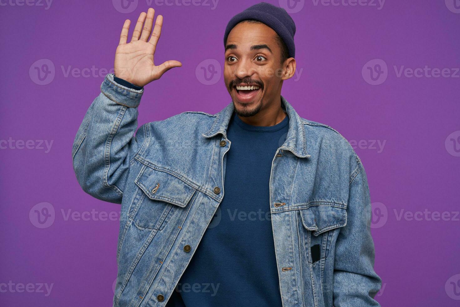 Happy young pretty dark skinned bearded brunette male rojoicing while meeting friend and raising palm in hello gesture, smiling happily while posing over purple background photo