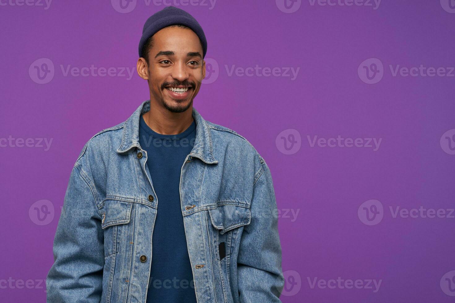 Cheerful young attractive bearded brunette man with dark skin looking positively aside with charming smile, wearing blue cap, pullover and jeans coat while posing over purple background photo