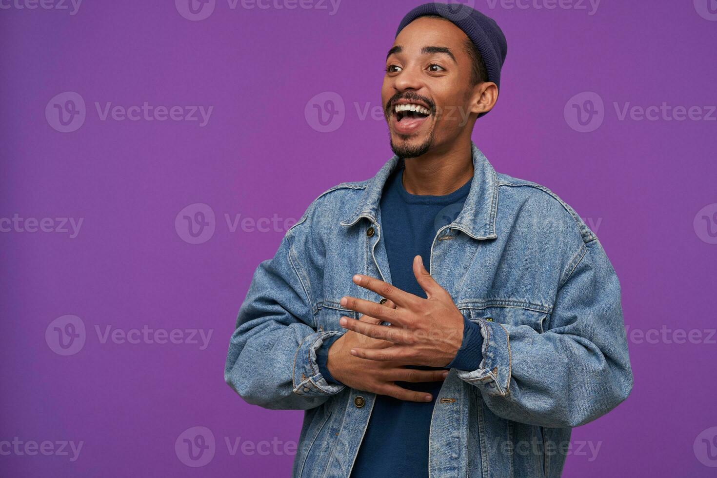 Joyous young handsome dark skinned bearded brunette male smiling joyfully and looking happily aside, dressed in casual clothes while posing over purple background photo