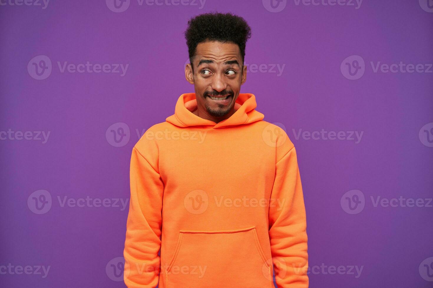 Studio shot of young lovely bearded dark skinned curly brunette guy looking positively aside with confused grimace, keeping hands down while standing over purple background photo