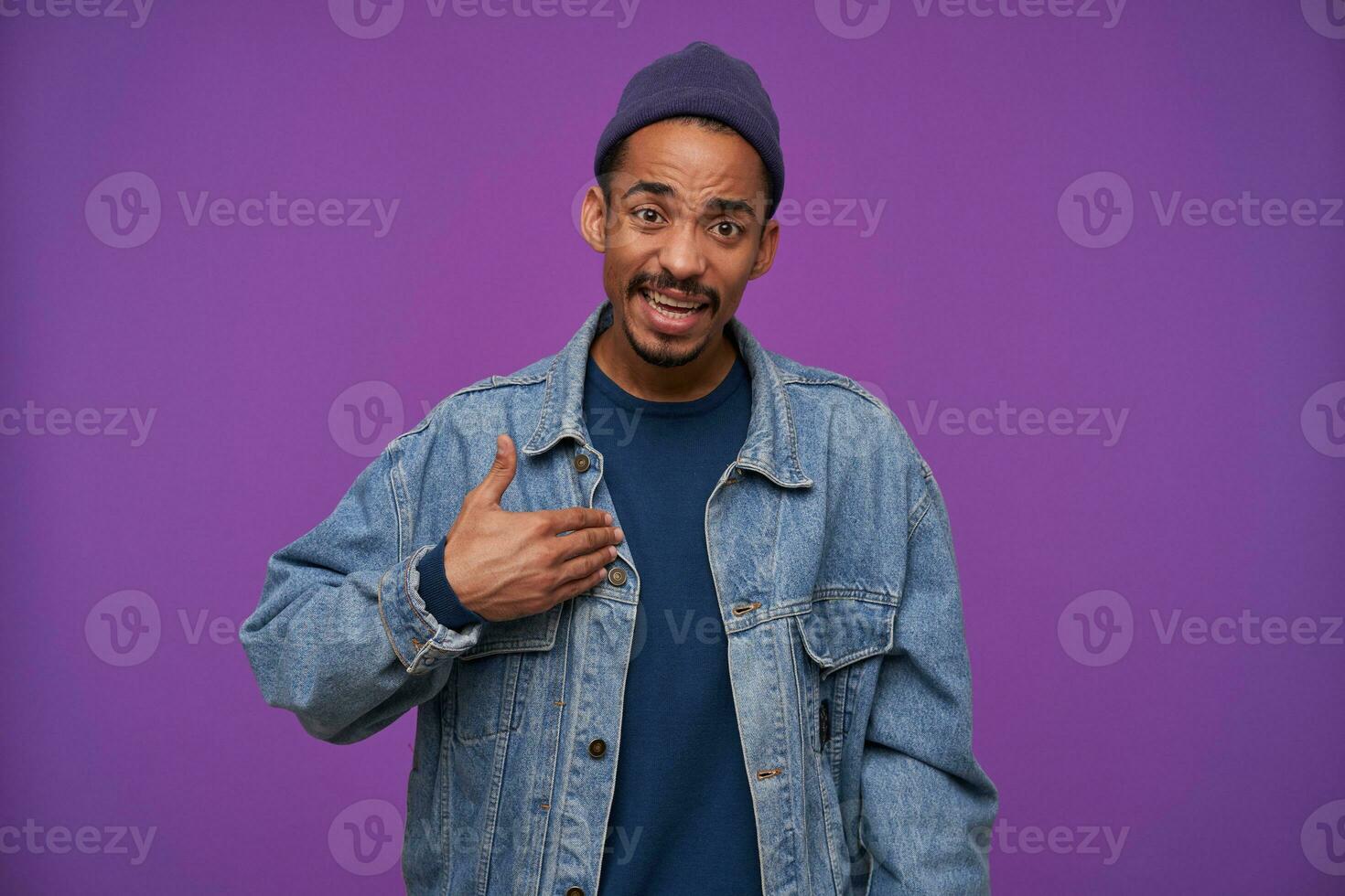 Puzzled young attractive dark skinned bearded brown haired man showing on himself with raised palm while looking confusedly to camera, standing against purple background photo