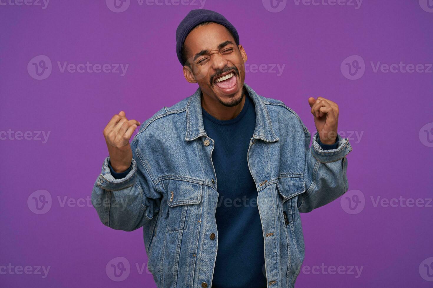 Portrait of pretty young happy dark skinned bearded brunette man keeping eyes closed while raising happily his fists, standing over purple background in casual clothes photo