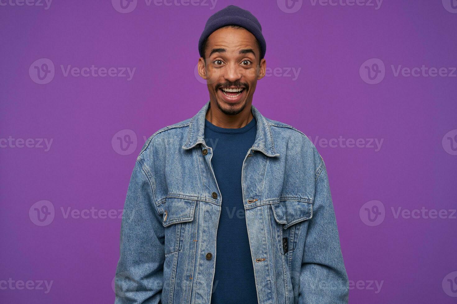 Indoor shot of young joyful dark skinned male with beard looking at camera with excited face, raising happily eyebrows while smiling widely, posing over purple background photo