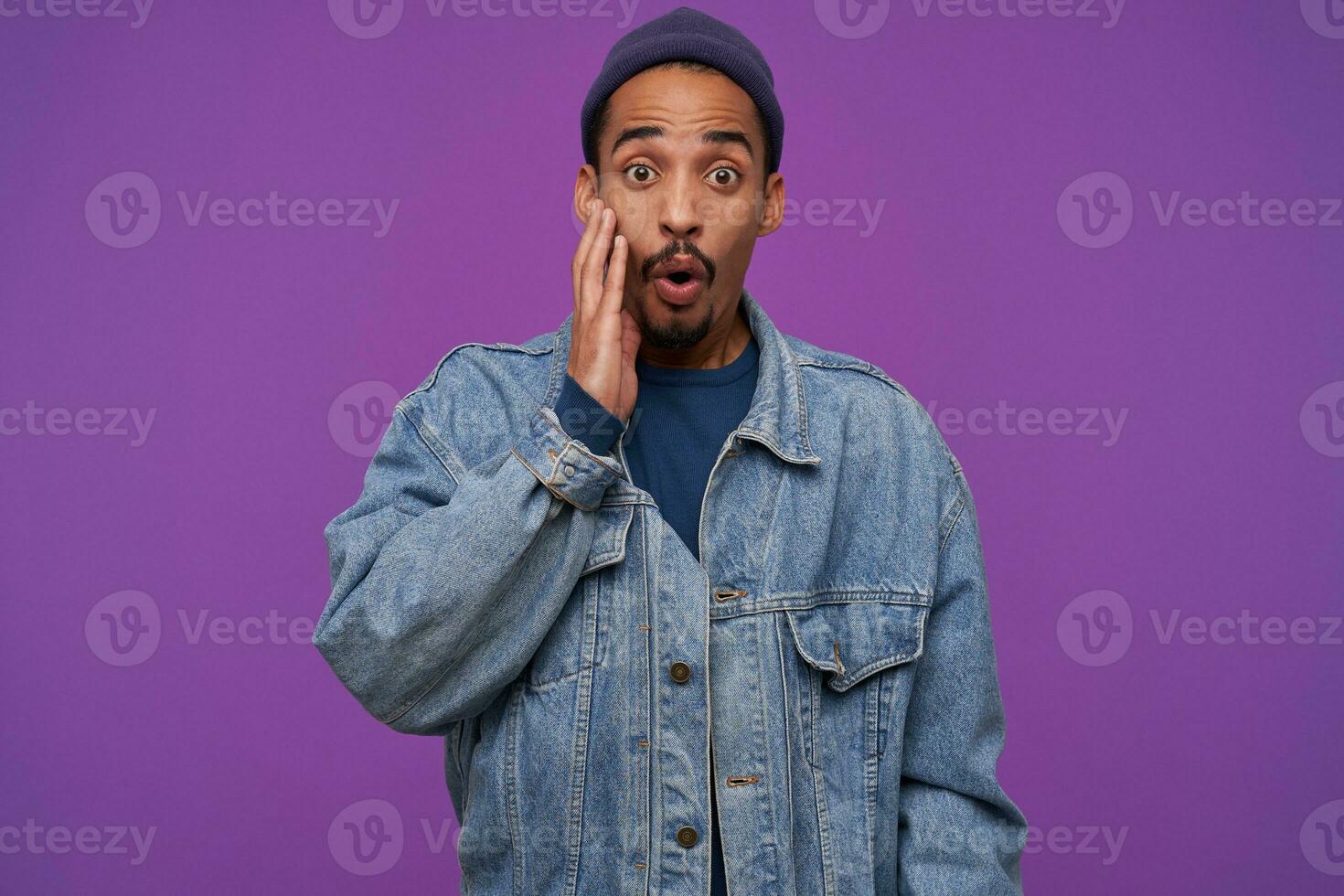 Astonished young dark skinned bearded brunette guy rounding his eyes while looking amazedly at camera, holding hand on his cheek while posing over purple background photo