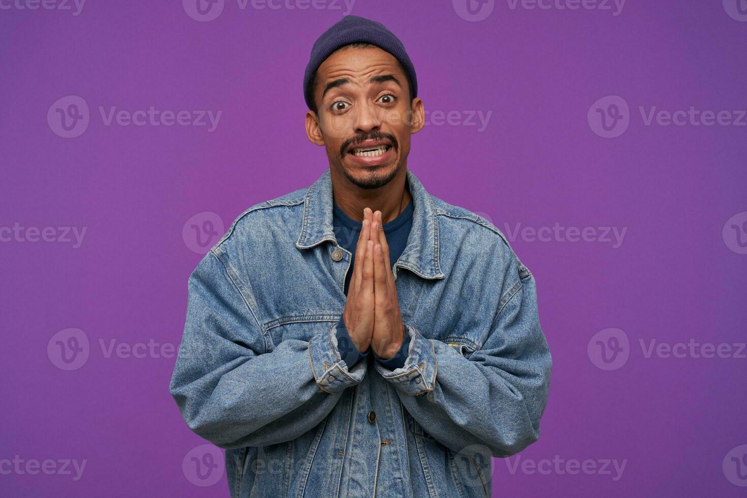 Indoor photo of young dark skinned bearded brunette man raising hands in praying gesture while posing over purple background, wrinkling forehead and showing teeth while looking at camera