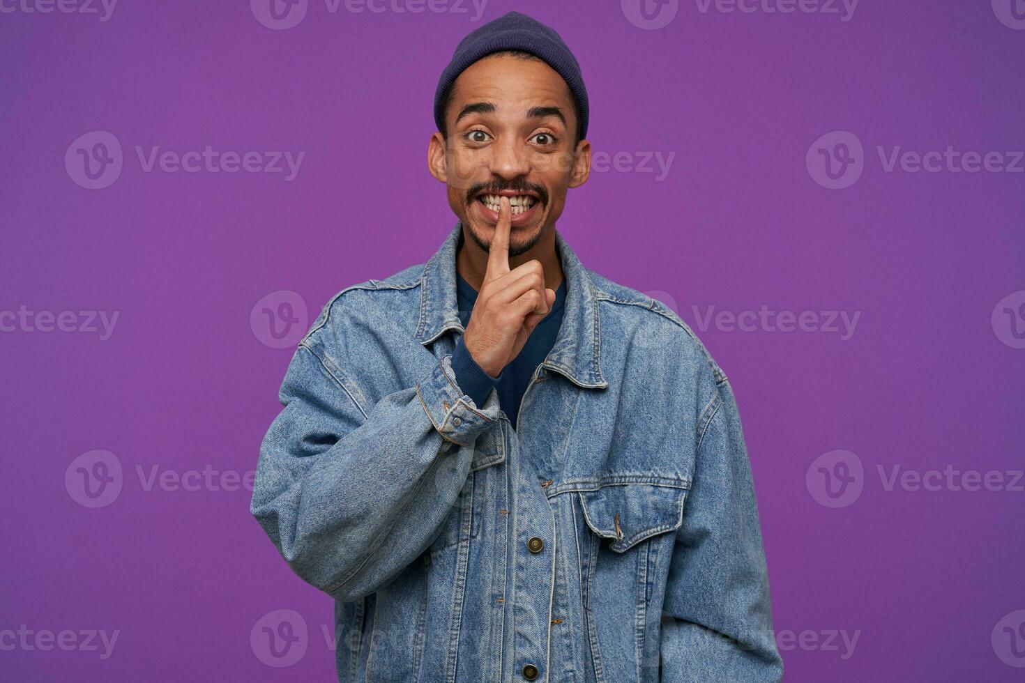 Indoor photo of young pretty bearded brunette male holding forefinger on his lips while looking at camera, calling to keep secret, standing over purple background