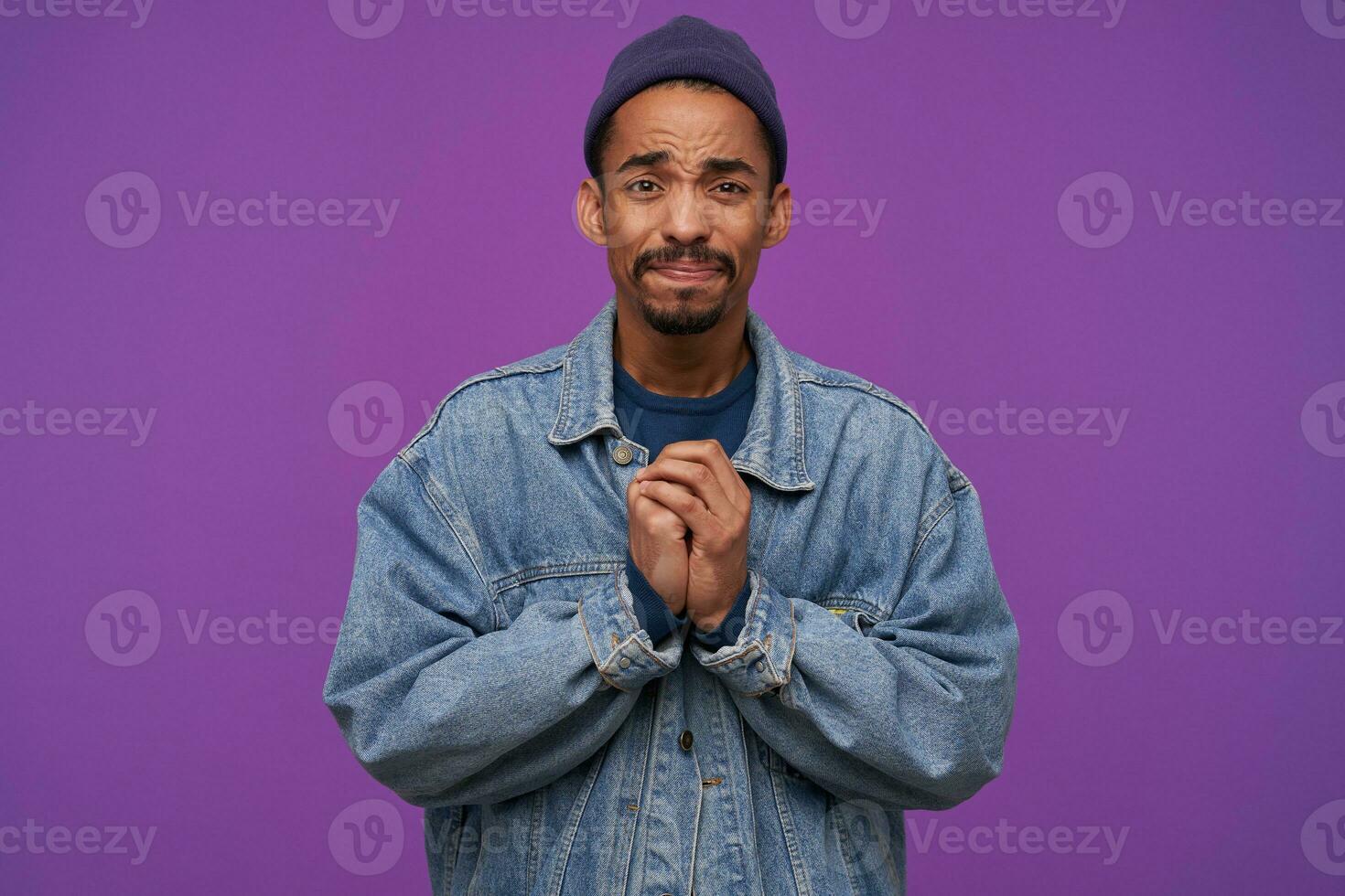 Sad young attractive bearded dark haired male looking worringly at camera and folding raised hands, frowning his upset face while standing over purple background photo