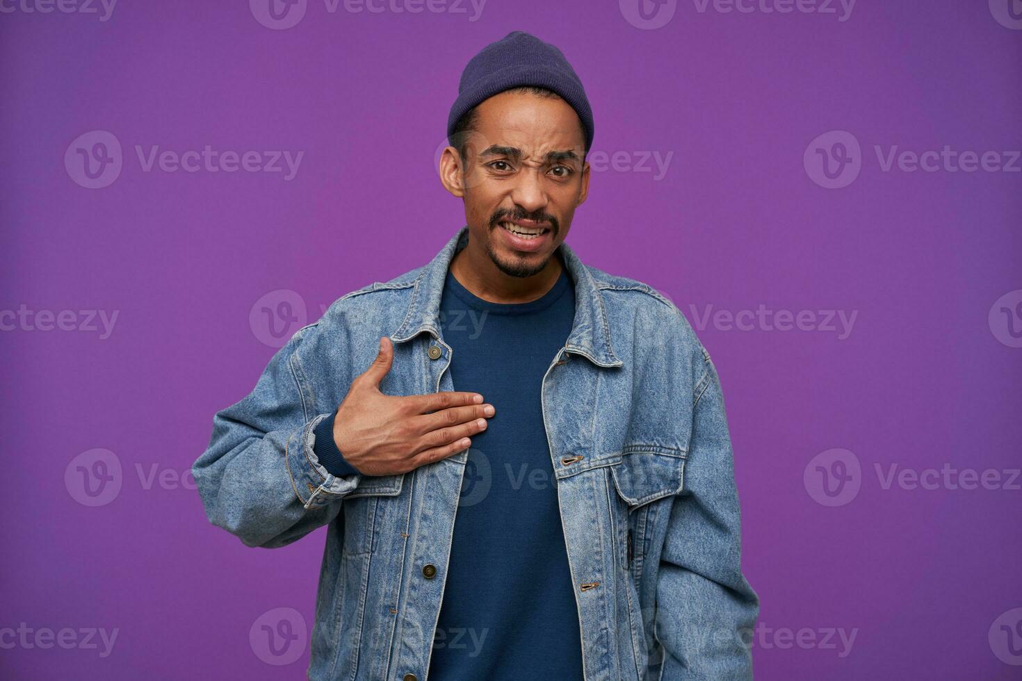 Confused young dark skinned bearded brunette guy frowning his eyebrows while looking at camera with puzzled face, dressed in casual wear while posing over purple background photo