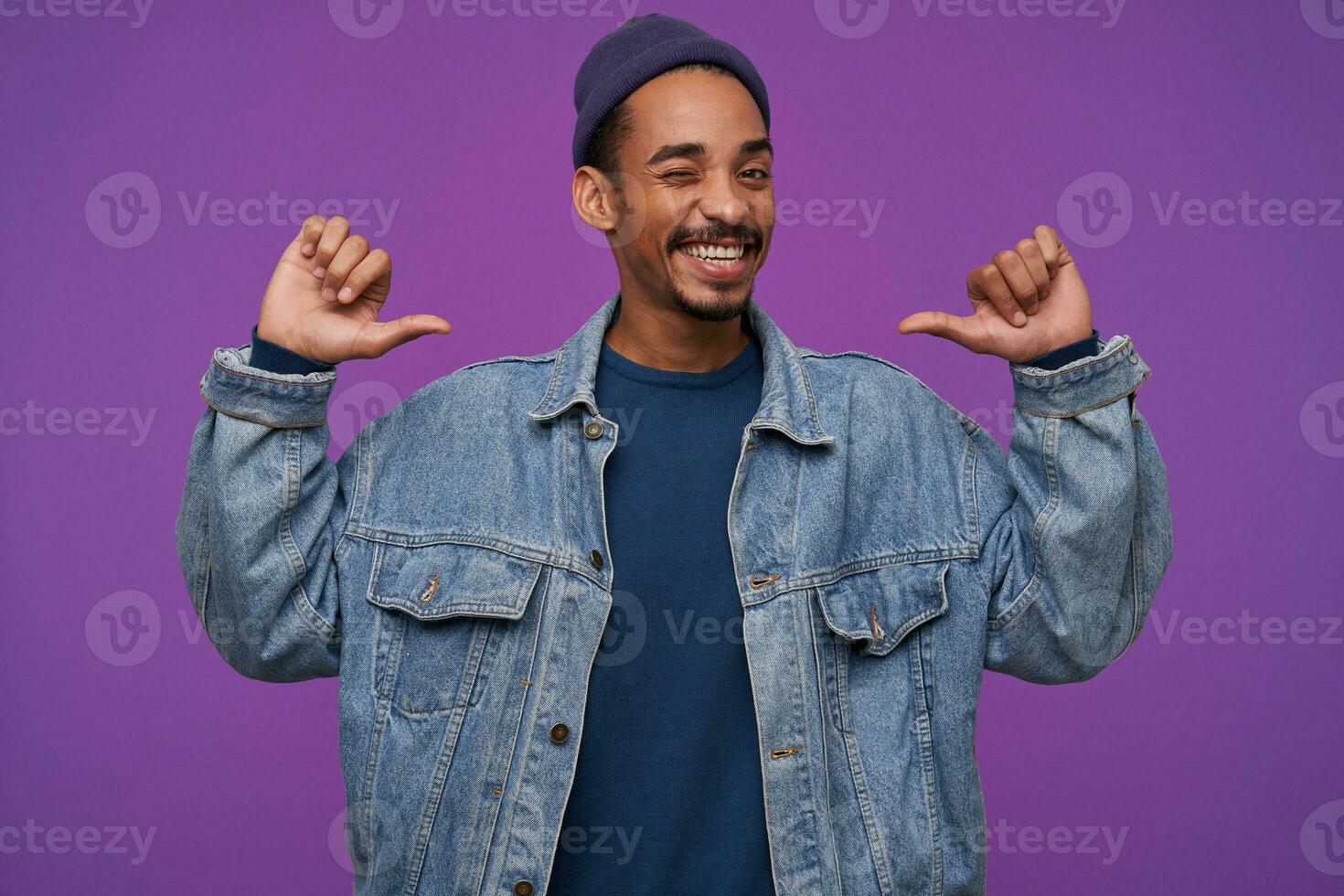 Cheerful young handsome bearded brunette guy with dark skin showing on himself with raised hands and smiling self-confidently at camera, isolated over purple background photo