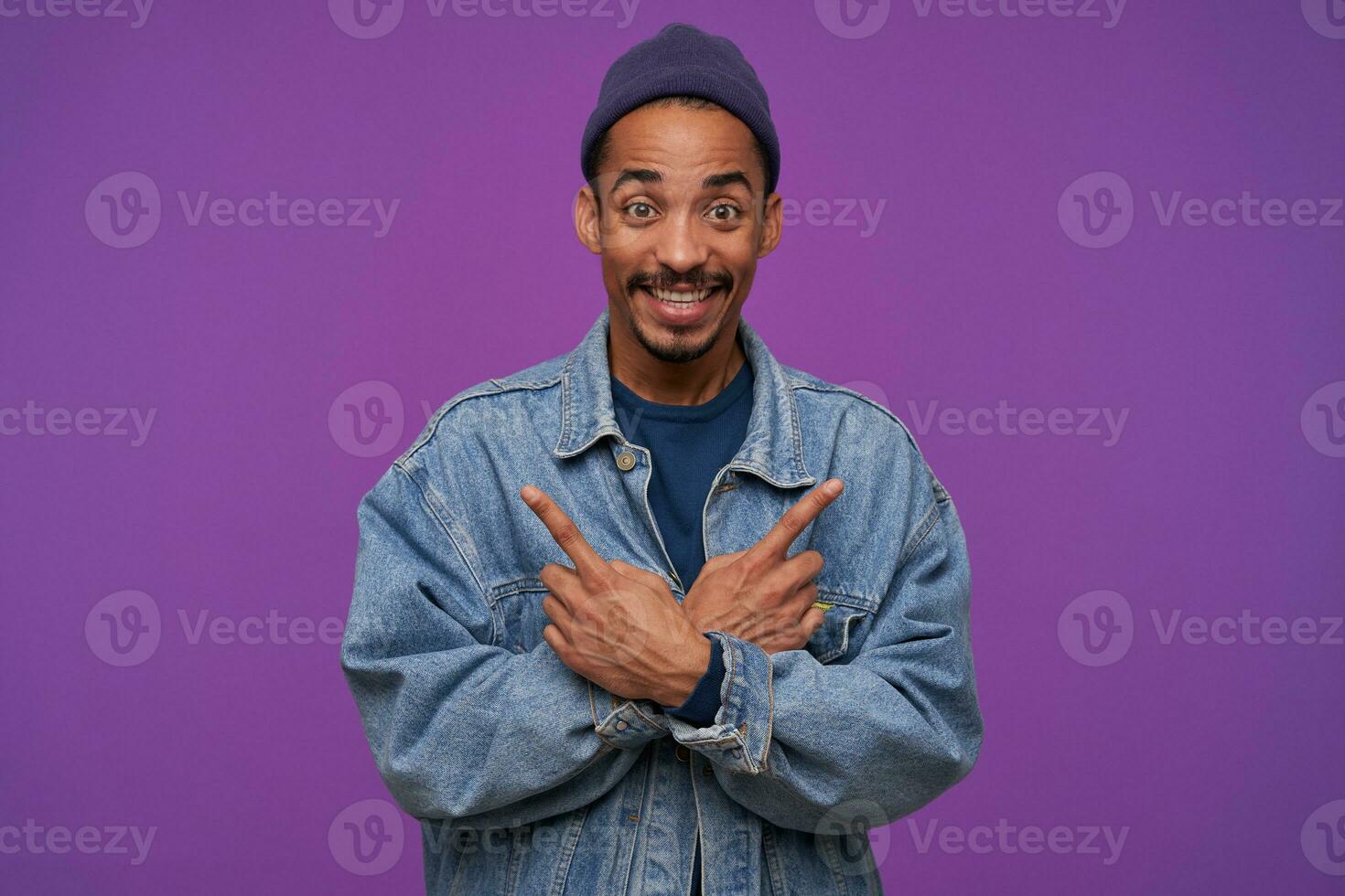 Indoor photo of cheerful young handsome dark skinned bearded man smiling widely while showing with forefingers in different directions, standing over purple background