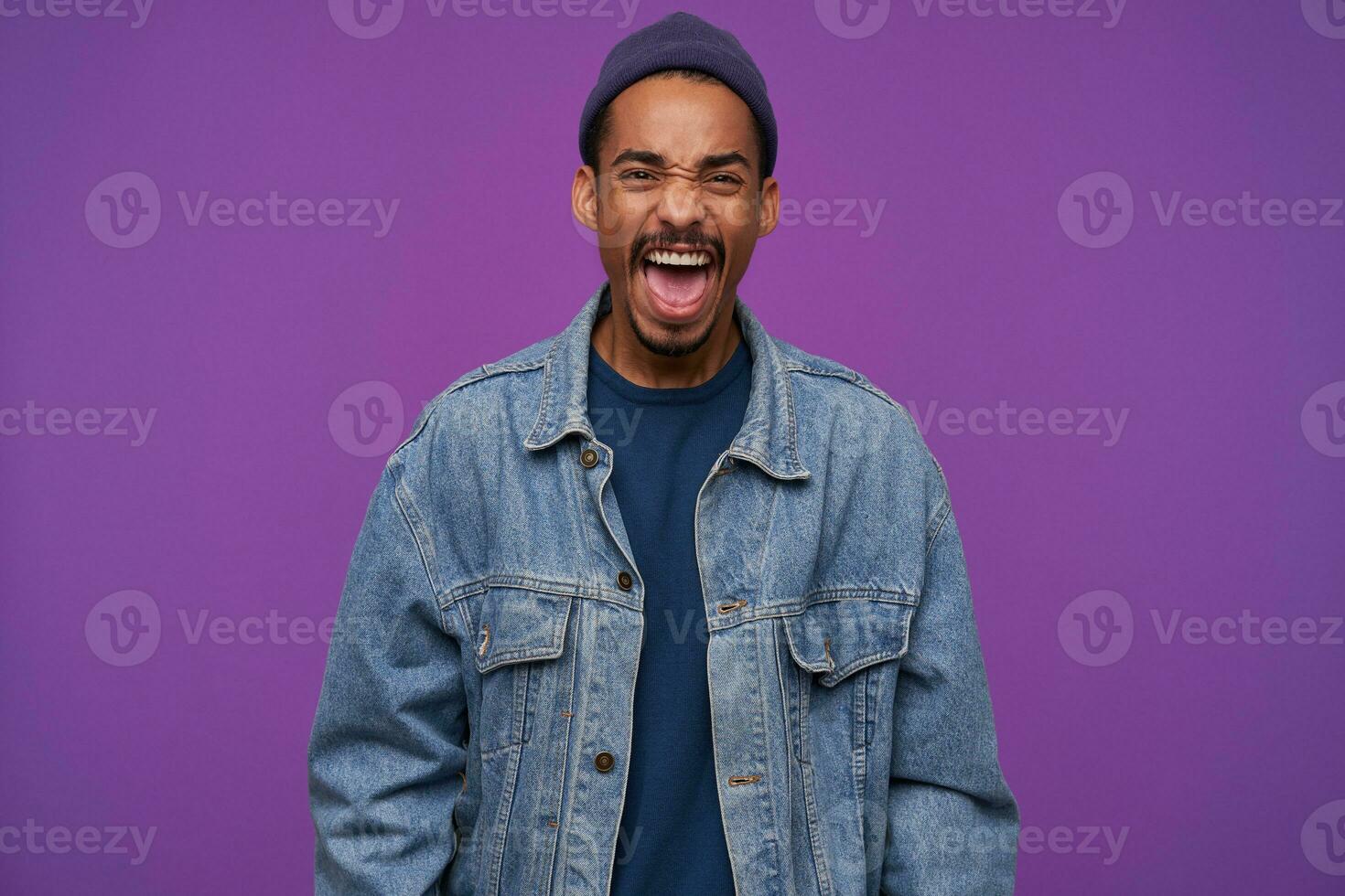 Indoor photo of young angry dark skinned brunette male with beard frowning his face while screaming fiercely with wide mouth opened, isolated over purple background